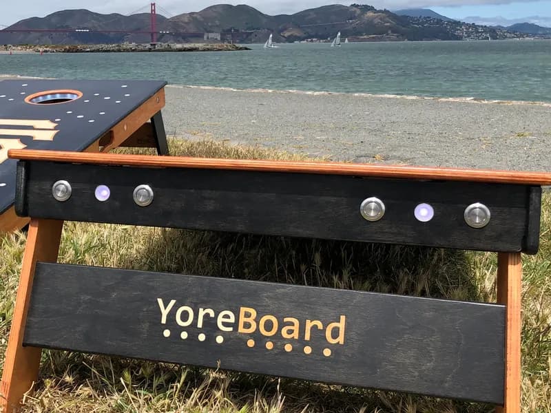 Detailed shot of YoreBoard cornhole boardss sides and backs, highlighting its functional integrated LED scoring buttons and hand-stained branding, with the Golden Gate Bridge in the background.