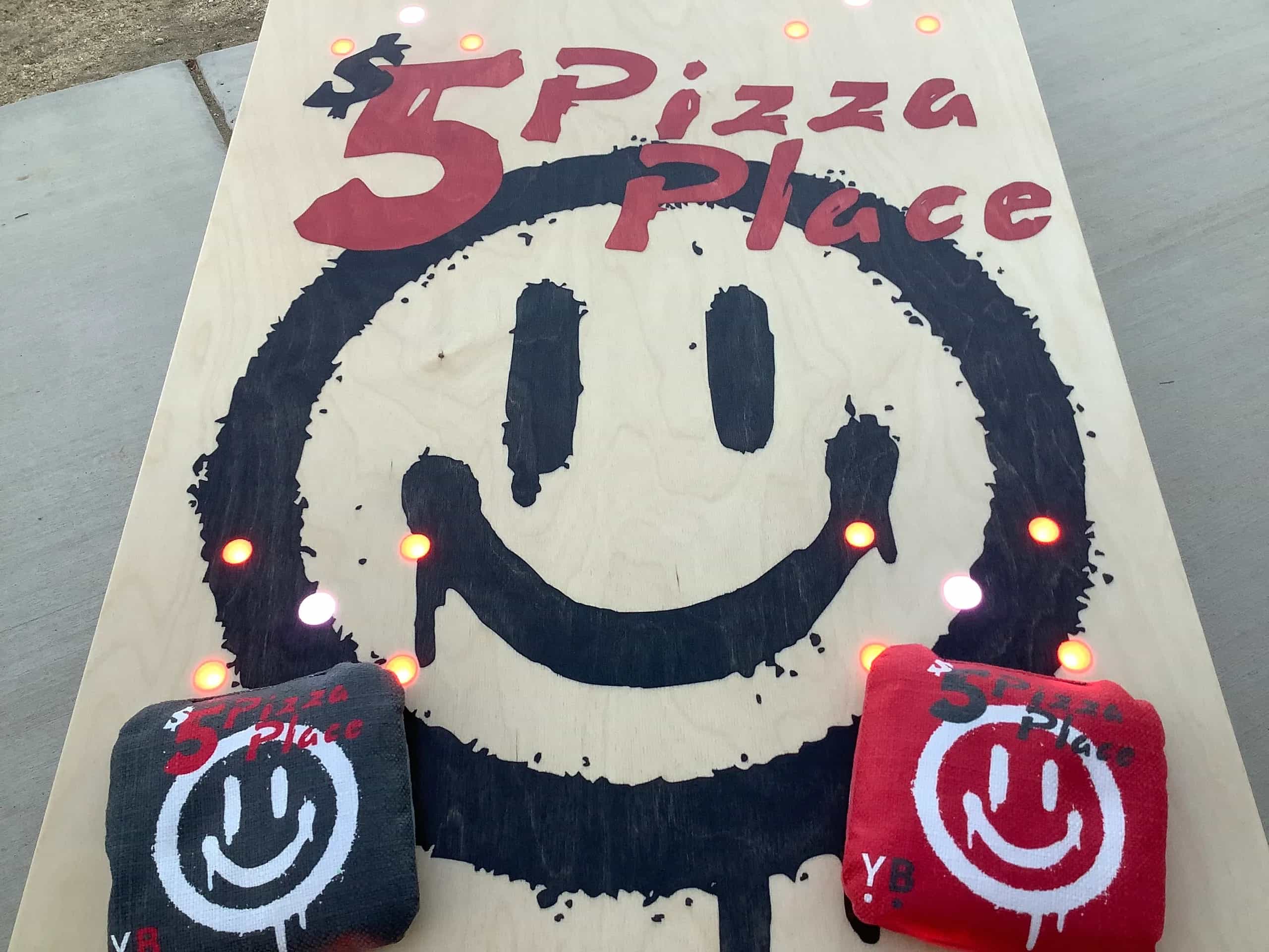 A close-up view of a YoreBoard cornhole board, featuring a hand-stained '5 Pizza Place' logo in red and a large, dripping black smiley face.