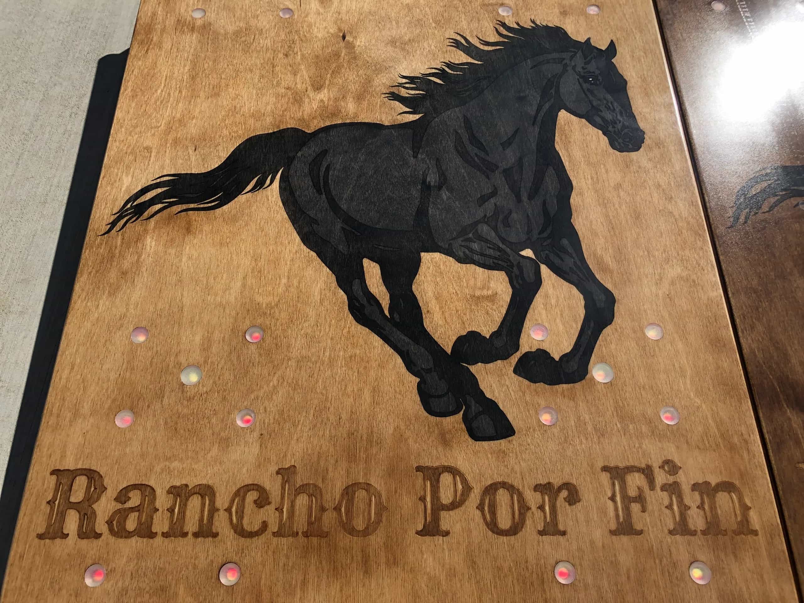 Close-up of a YoreBoard cornhole board with a hand-stained running horse graphic and 'Rancho Por Fin' text, with integrated LED scoring.