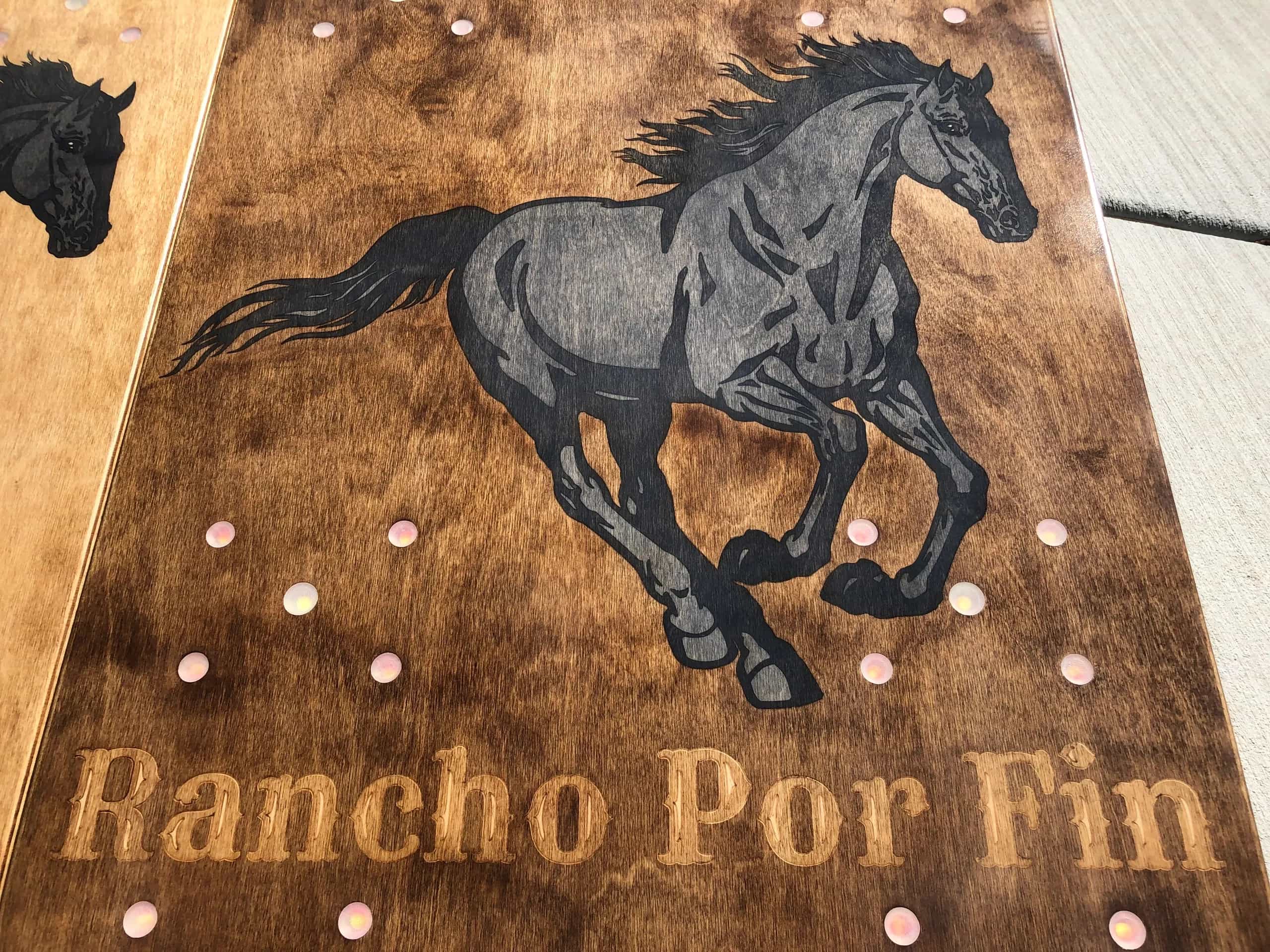 Close-up of a YoreBoard cornhole board with a hand-stained running horse graphic and 'Rancho Por Fin' text, with integrated LED scoring.