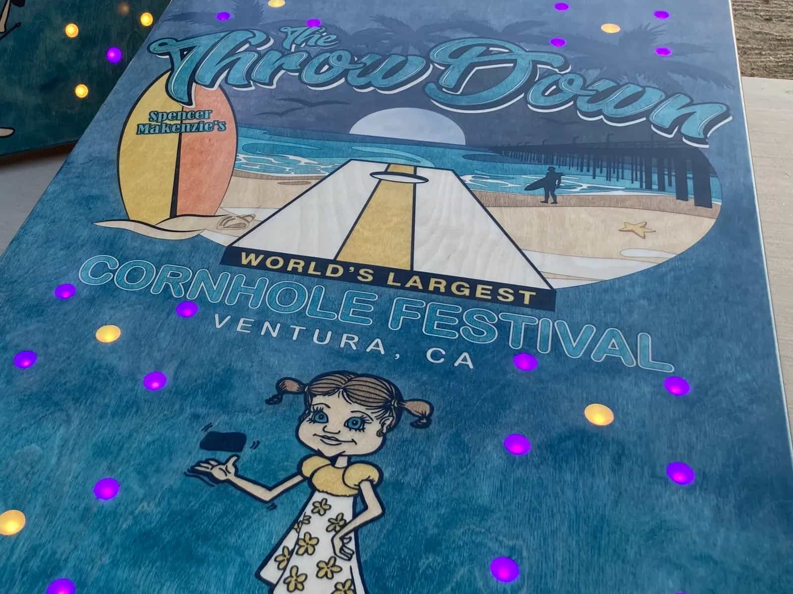 Detailed shot of a YoreBoard cornhole board highlighting its custom hand-stained festival artwork with a playful beach theme, illuminated by integrated LED scoring, emphasizing the board's vibrant design and features.