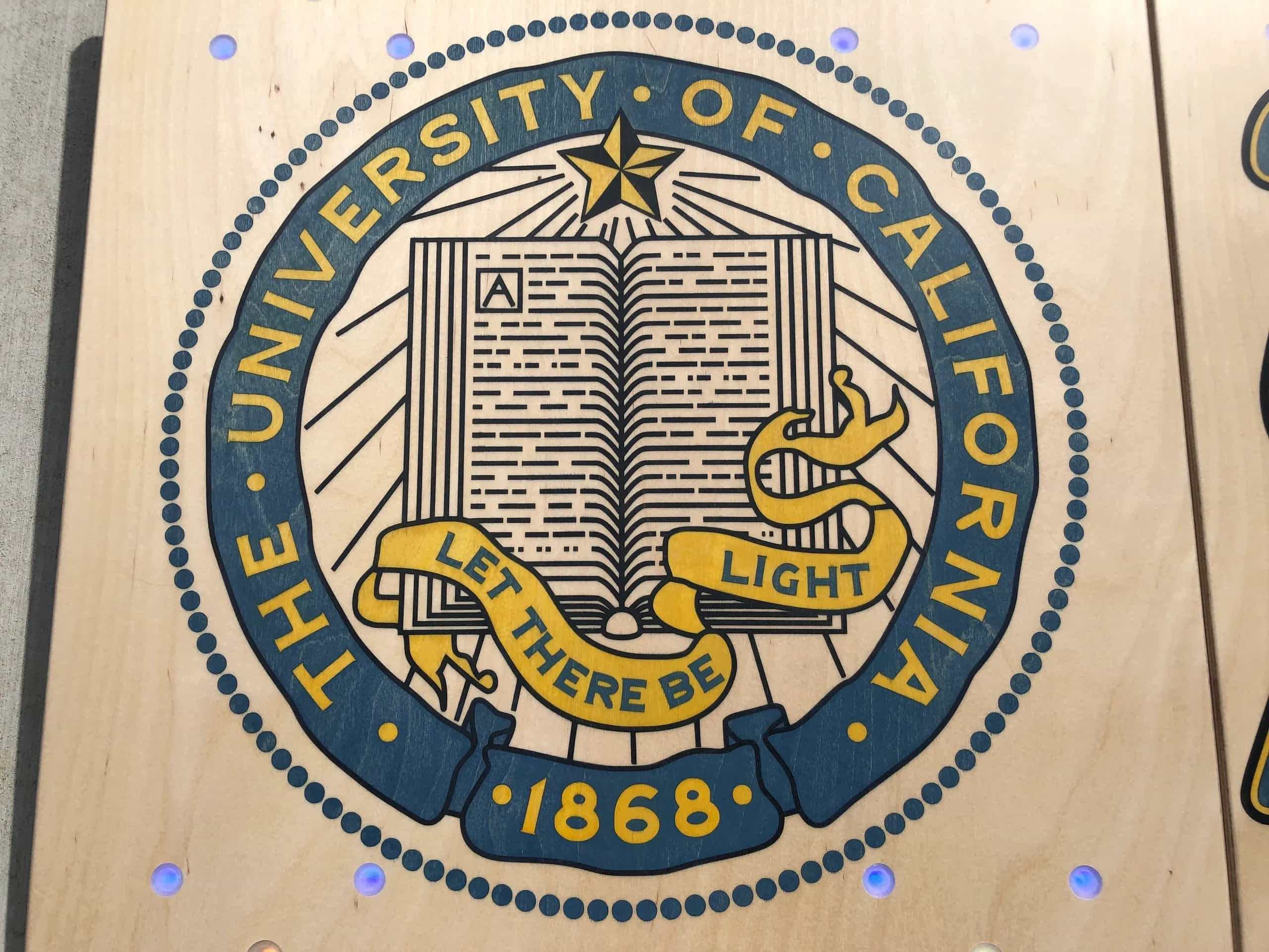A close-up view of a YoreBoard cornhole board surface, showcasing a large, hand-stained University of California seal in blue and gold, featuring an open book and the motto 'LET THERE BE LIGHT'.