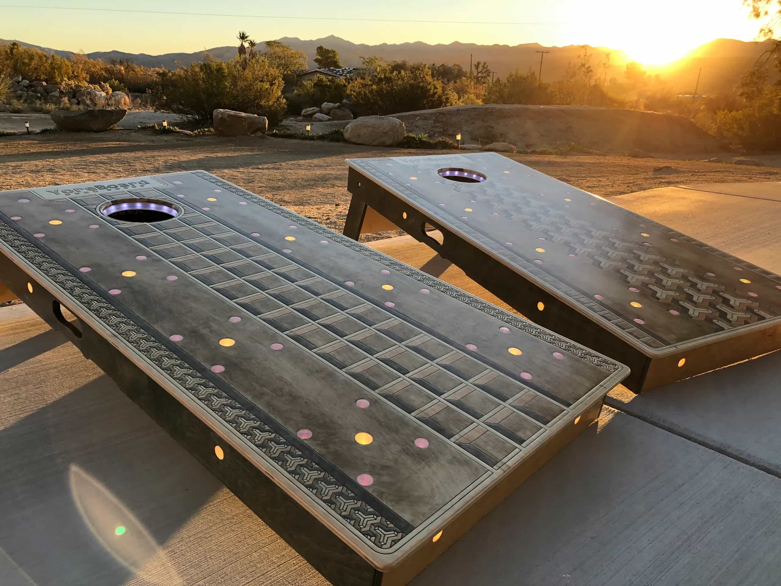 A pair of YoreBoard cornhole boards showcasing intricate carved designs and glowing embedded lights, captured during golden hour in a desert environment.