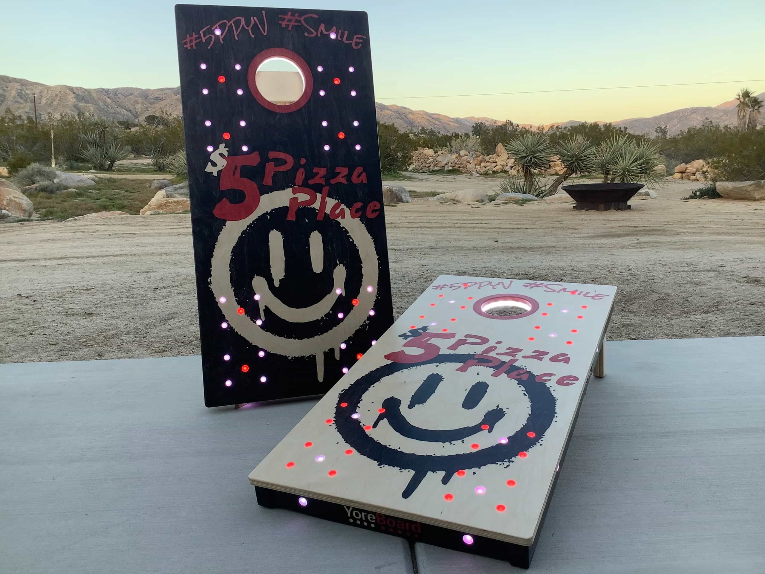 A pair of YoreBoard cornhole boards designed with a '5 Pizza Place' theme, including a dripping paint effect and glowing LED lights, ideal for brand promotion or events.