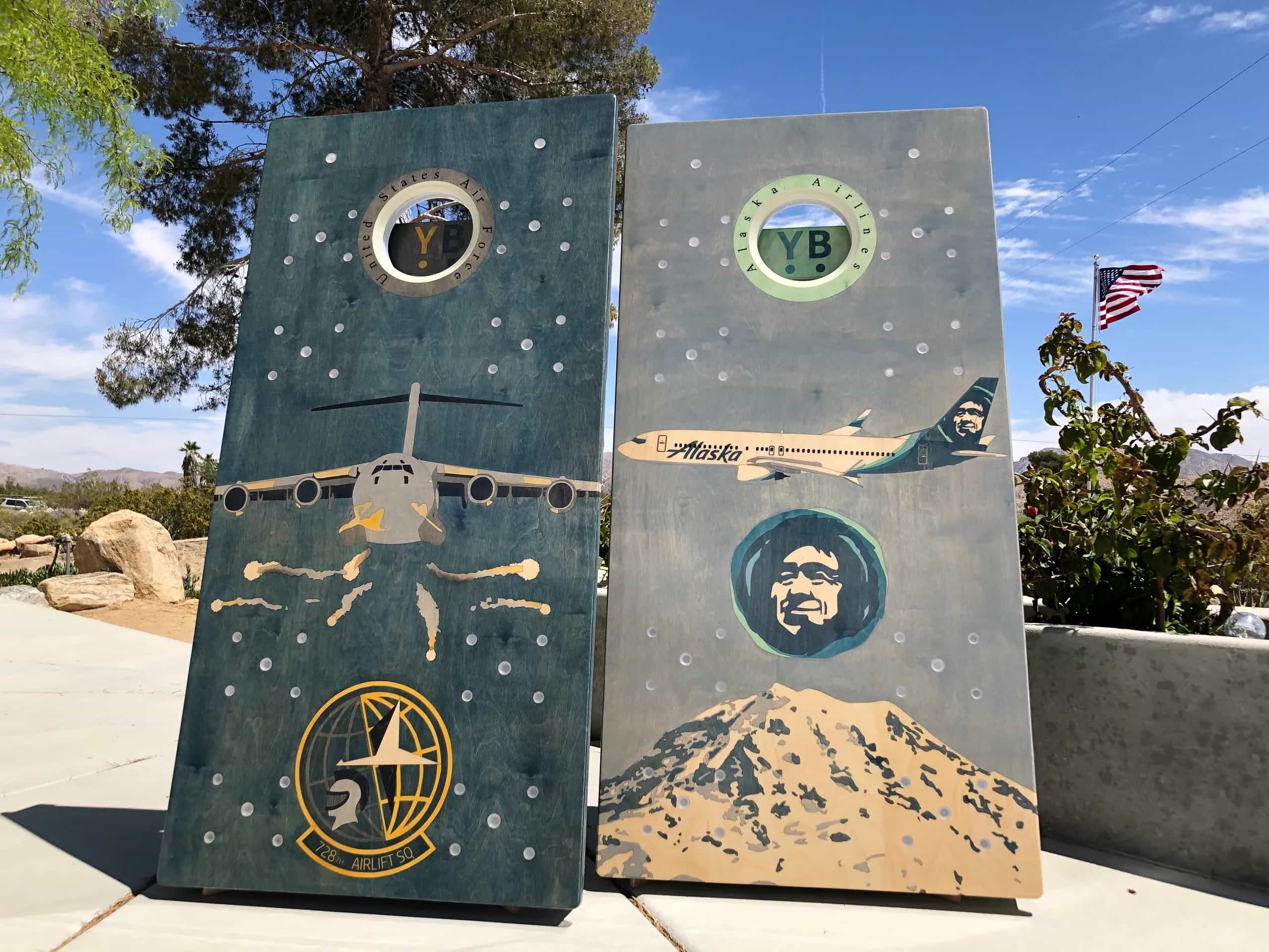 A pair of YoreBoard cornhole boards showcasing detailed hand-stained designs of military transport aircraft and a commercial passenger jet, alongside various aviation and geographical motifs, under a clear sky.