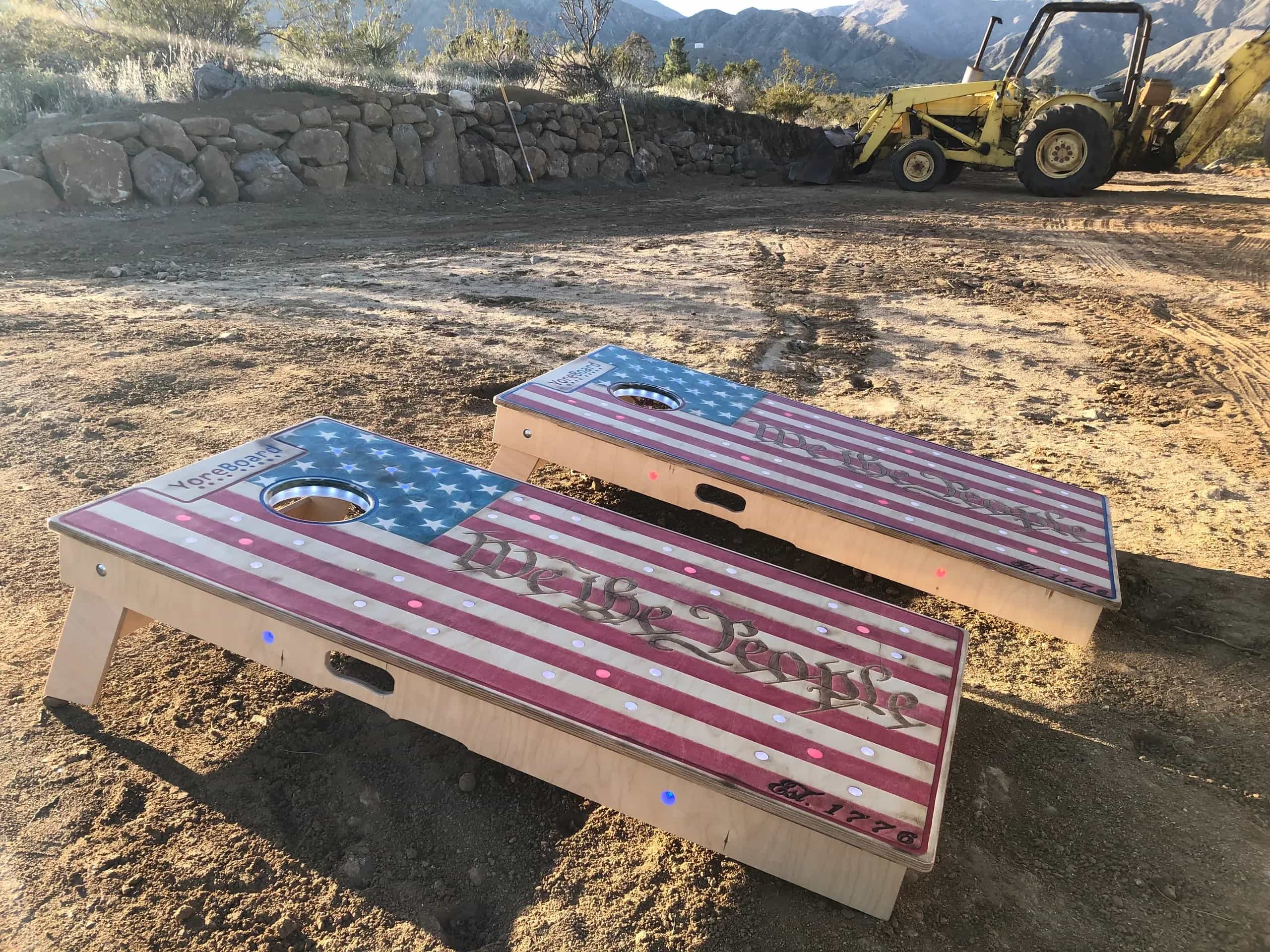 Two custom YoreBoard cornhole boards, each hand-stained with an American Flag design and 'We the People' text, featuring embedded LED lights, set on a dirt path outdoors with a yellow tractor and mountains in the background.