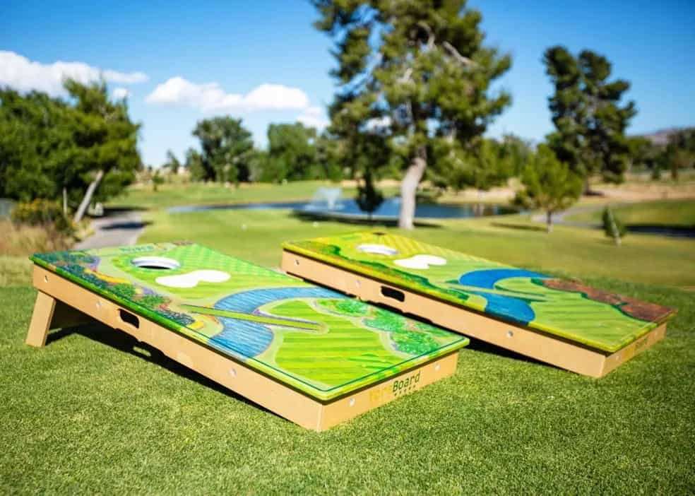 Two custom YoreBoard cornhole boards, each hand-stained with a vibrant golf course hole design featuring greens, fairways, and water hazards, set on lush green grass with a golf course and trees in the background under a sunny sky.