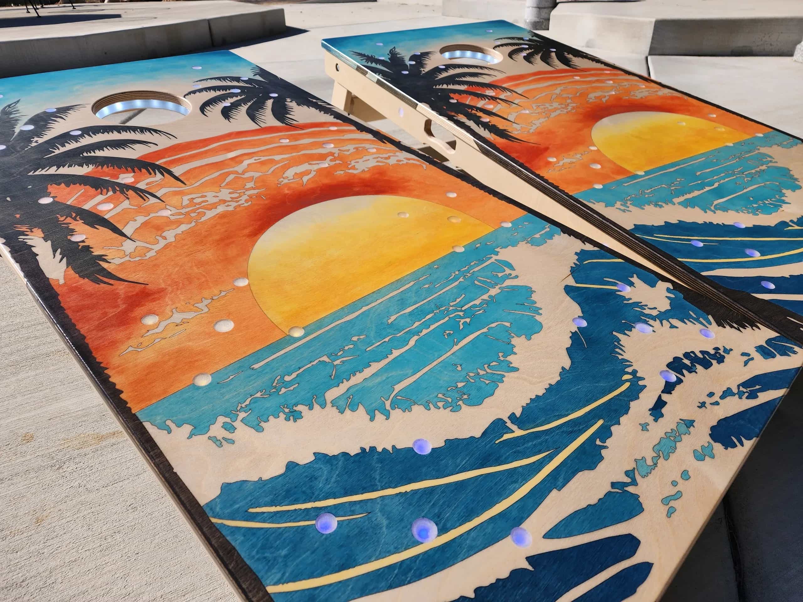 A pair of YoreBoard cornhole boards showcasing detailed hand-stained artwork of a tropical sunset over the ocean, with integrated lighting, designed for a relaxed, beach-themed atmosphere.
