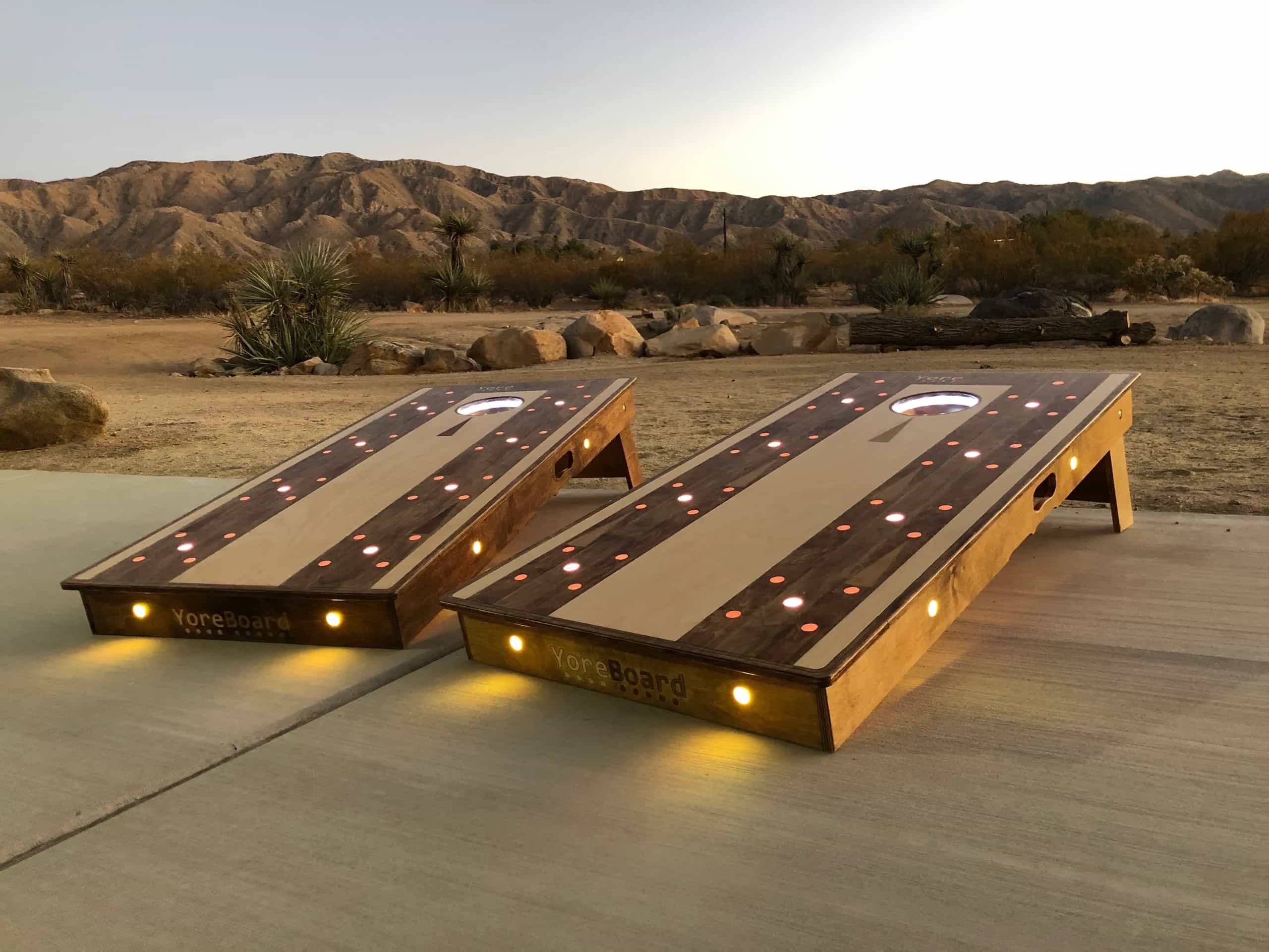 Two custom YoreBoard cornhole boards, each hand-stained to resemble a bowling alley lane with light and dark wood stripes and embedded warm yellow and orange LED lights, set outdoors on a paved surface with desert mountains and a sunset sky in the background.