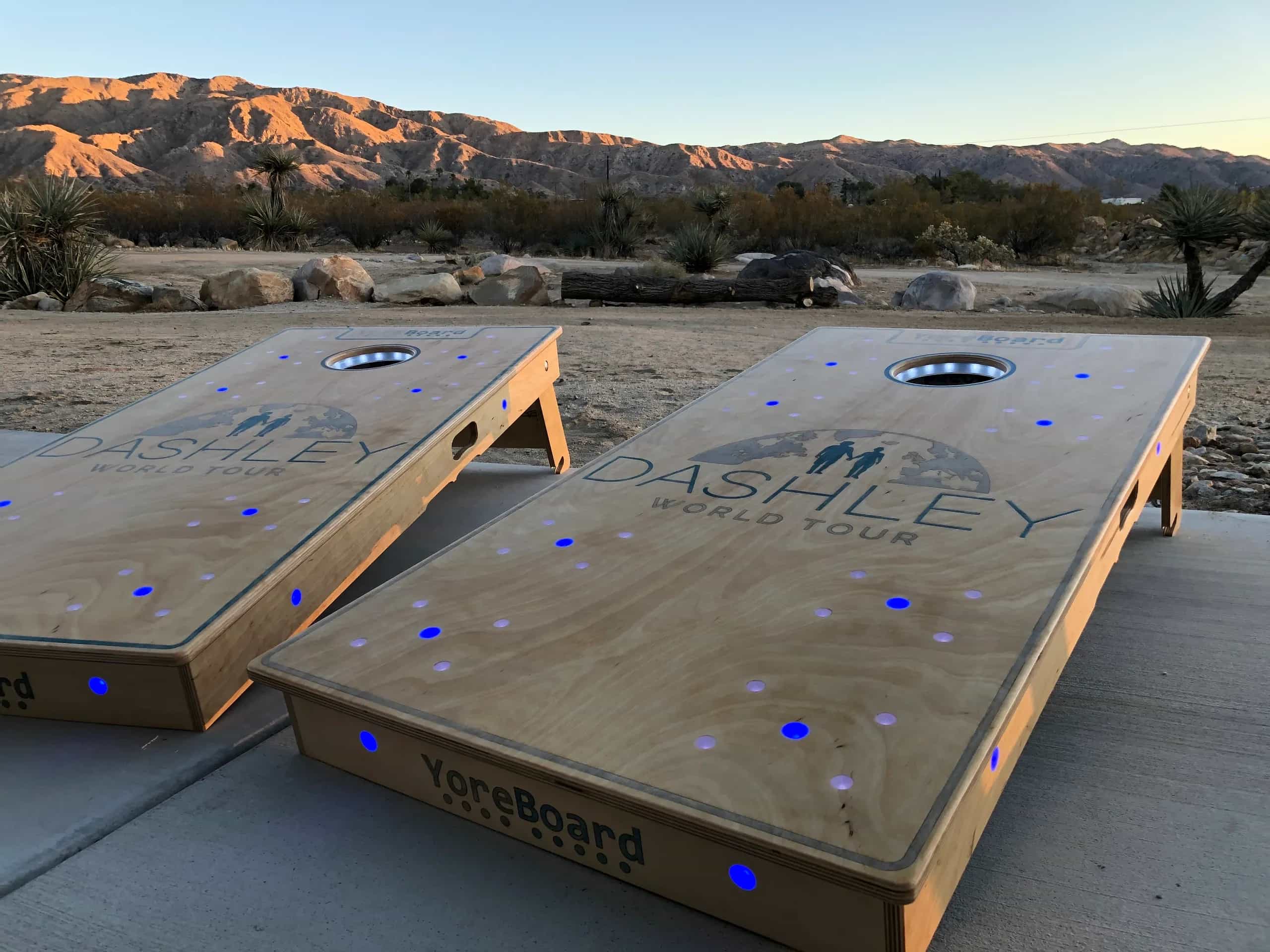 Two custom YoreBoard cornhole boards, each hand-stained with a 'DASHLEY World Tour' logo and embedded blue LED lights, set outdoors with desert mountains and vegetation in the background at sunset.