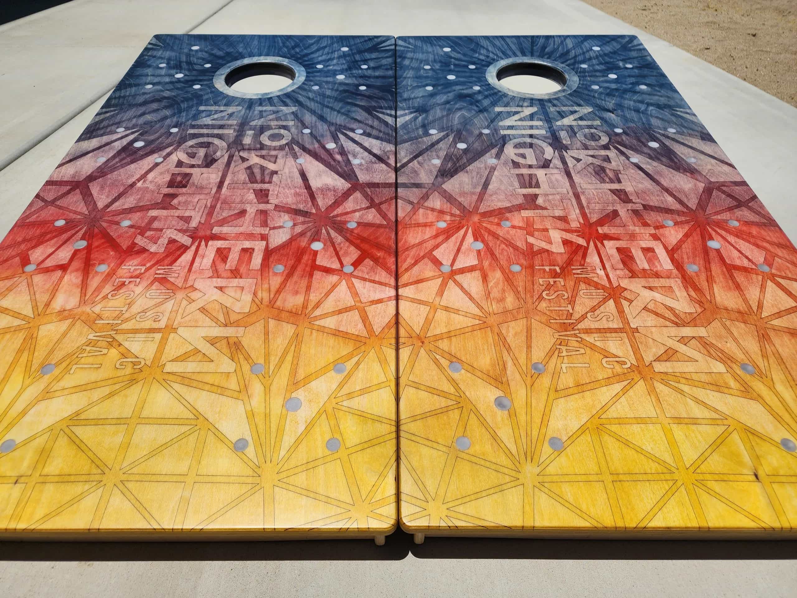 A pair of YoreBoard cornhole boards showcasing intricate, hand-stained artwork with a gradient color scheme and festival branding, designed for a lively event atmosphere.