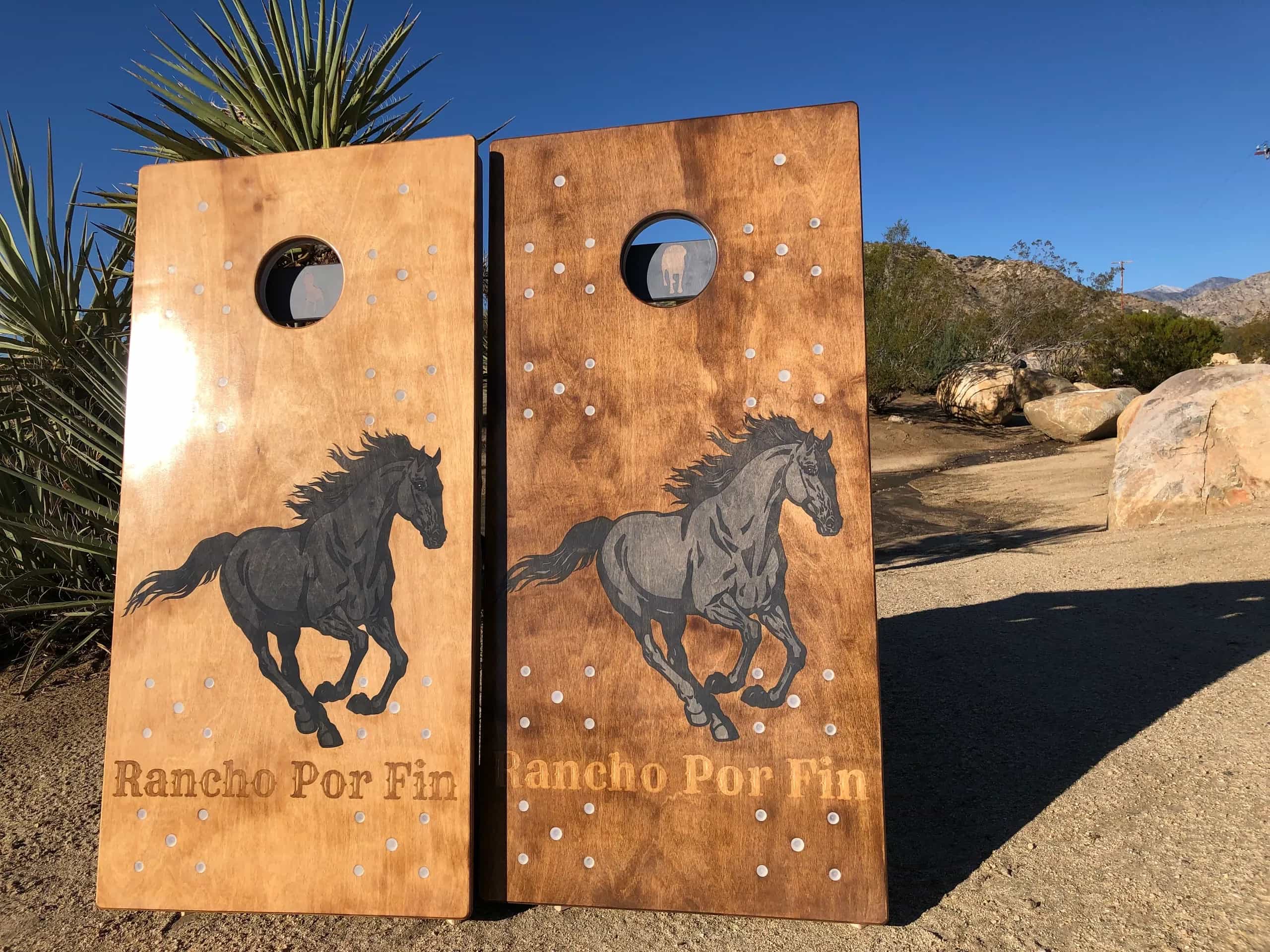 Two custom YoreBoard cornhole boards, one light-stained and one dark-stained, both featuring a hand-stained image of a running horse and 'Rancho Por Fin' text, with small embedded light dots. They are set outdoors near desert plants and rocks under a clear sky.