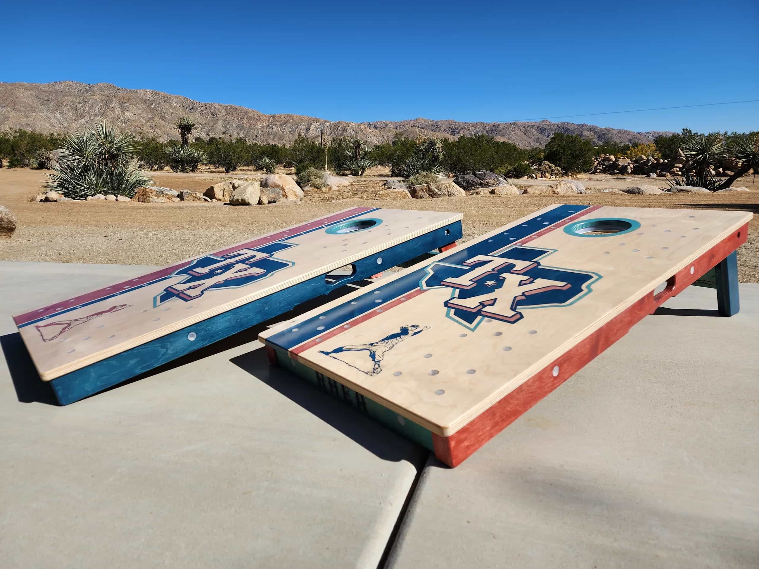 Two custom YoreBoard cornhole boards, one light-stained and one with red and blue accents, both featuring a hand-stained Texas Rangers 'TX' logo and a baseball player silhouette, with small embedded light dots. They are set outdoors with desert plants, rocks, and mountains in the background under a clear sky.