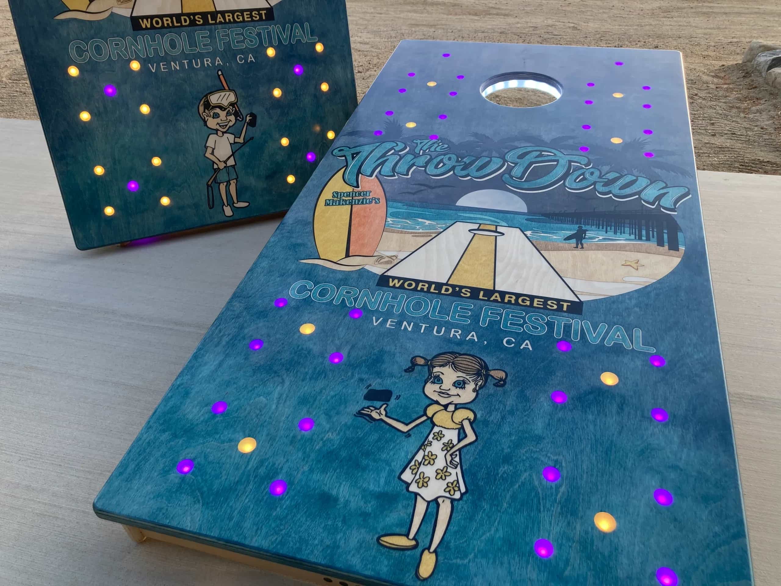 A pair of YoreBoard cornhole boards showcasing vibrant, hand-stained festival artwork with a playful design and glowing LED illumination, perfect for a large-scale cornhole event.