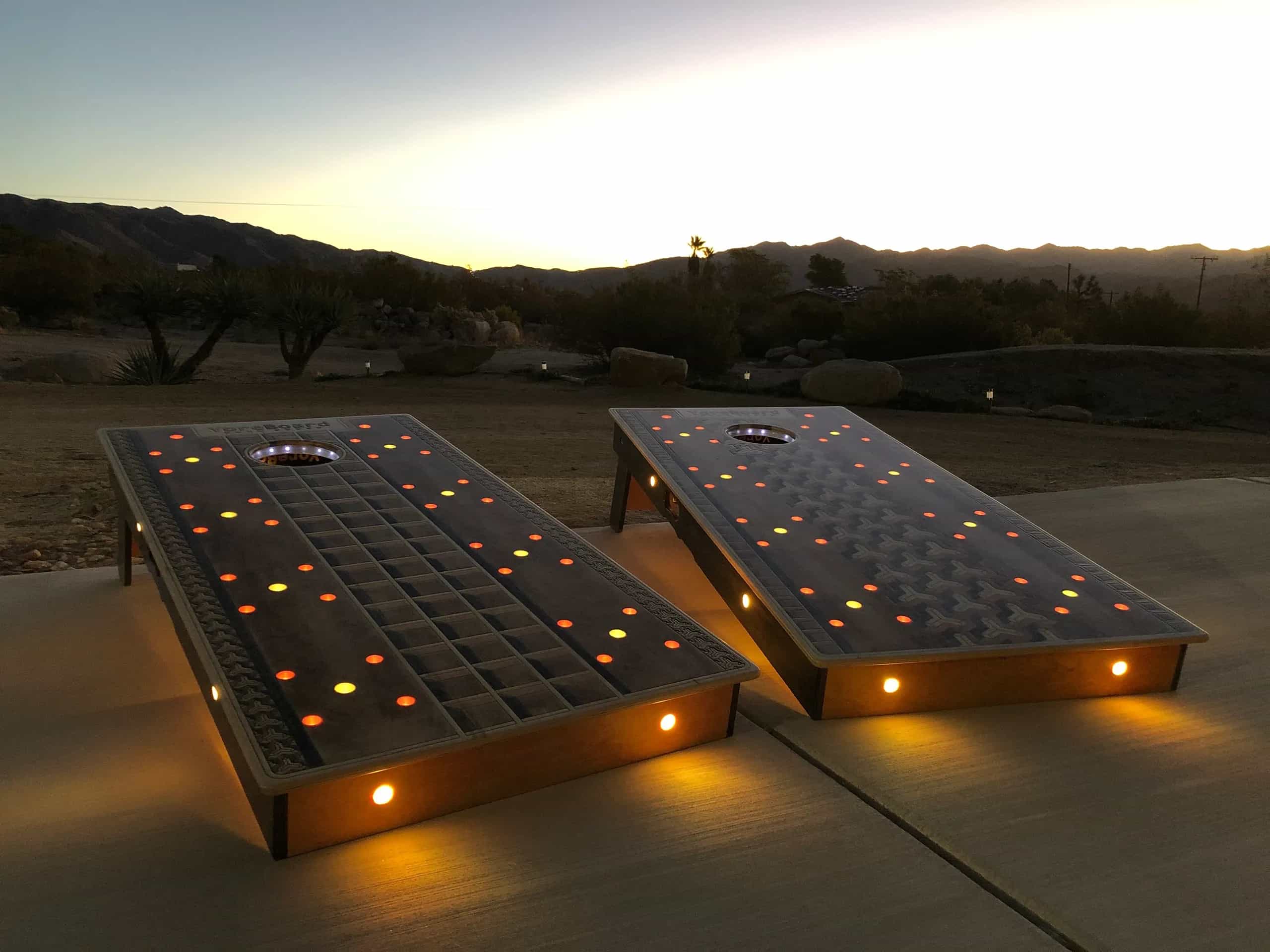 YoreBoard cornhole set lit up with colorful red and yellow LEDs, perfect for evening games, with a scenic sunset backdrop.
