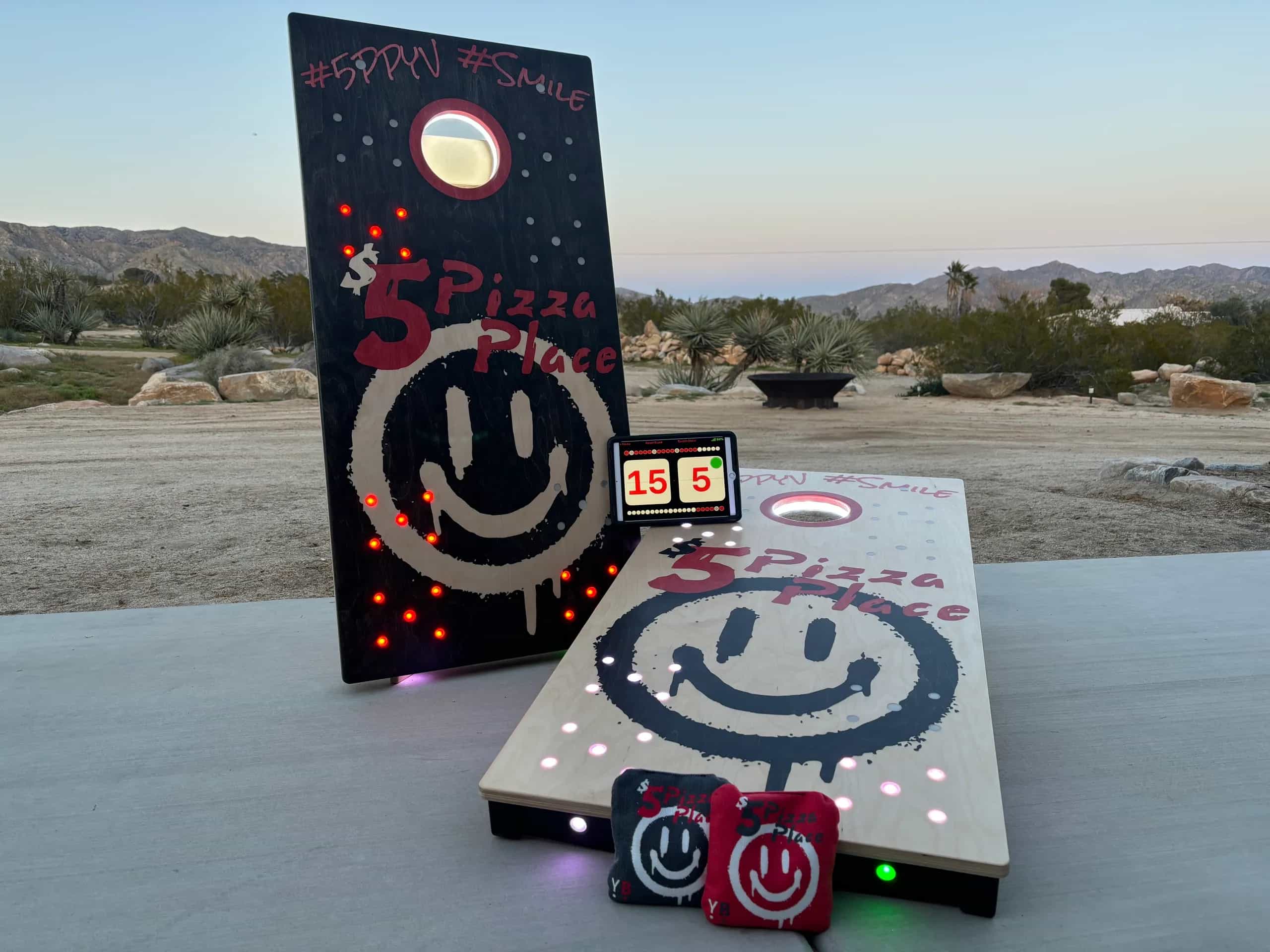 A detailed view of '5D Pizza Place' branded YoreBoard cornhole boards with active LED lighting, scorekeeping tablet, and matching cornhole bags ready for play.