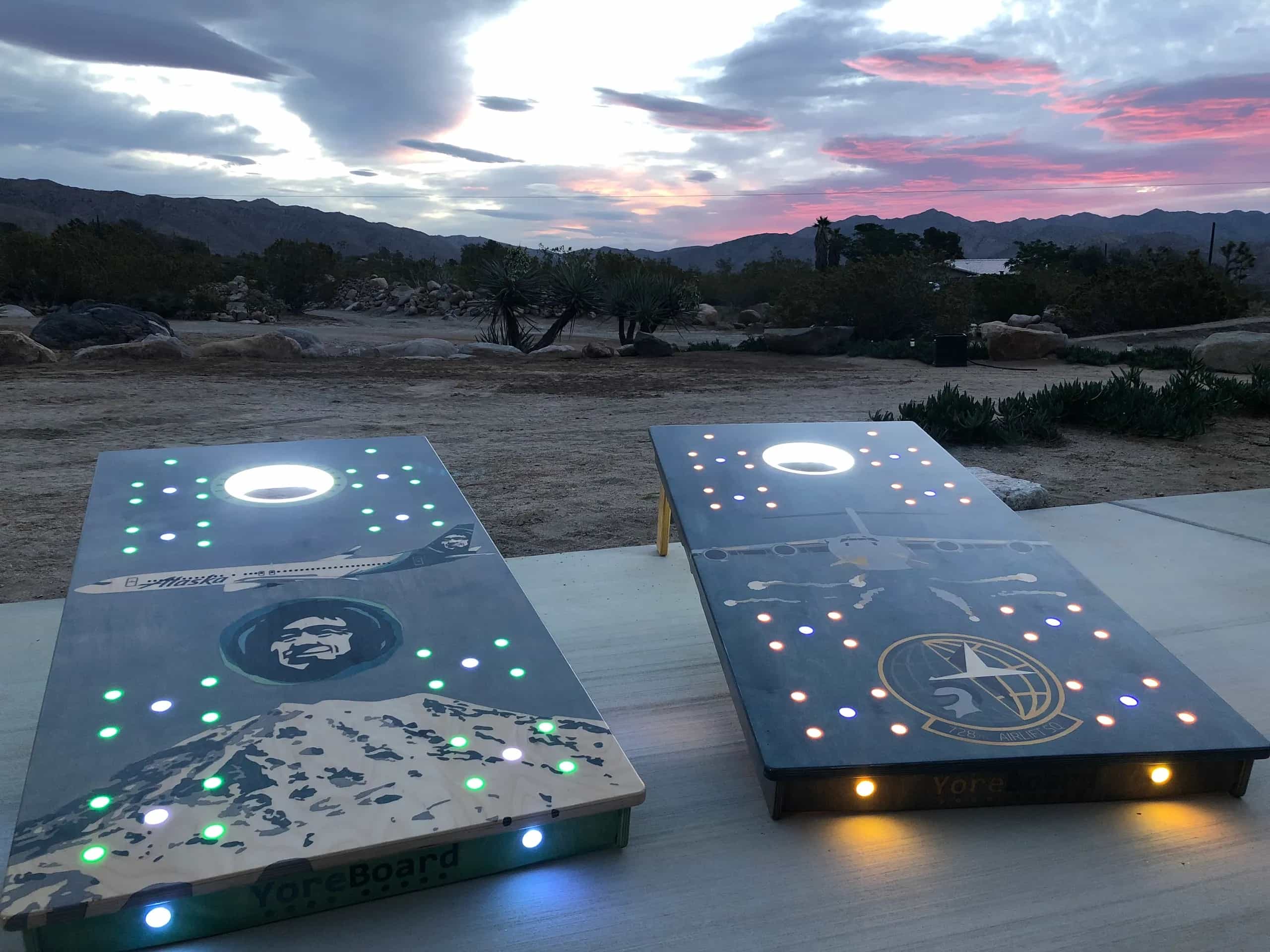 Personalized YoreBoard cornhole set featuring distinct airplane and portrait artwork with vibrant purple, green, and yellow LED lighting for evening play.