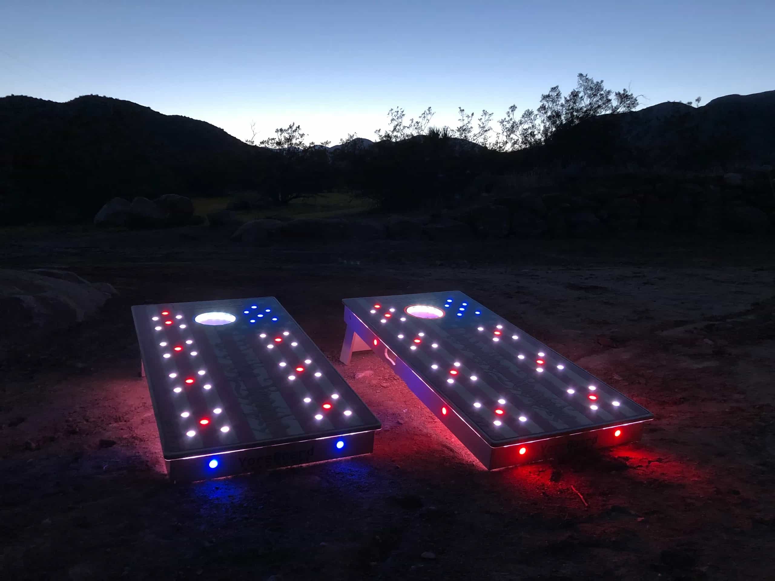 Two custom YoreBoard cornhole boards with an American flag design, illuminated by red, white, and blue Scoring LEDs at dusk.