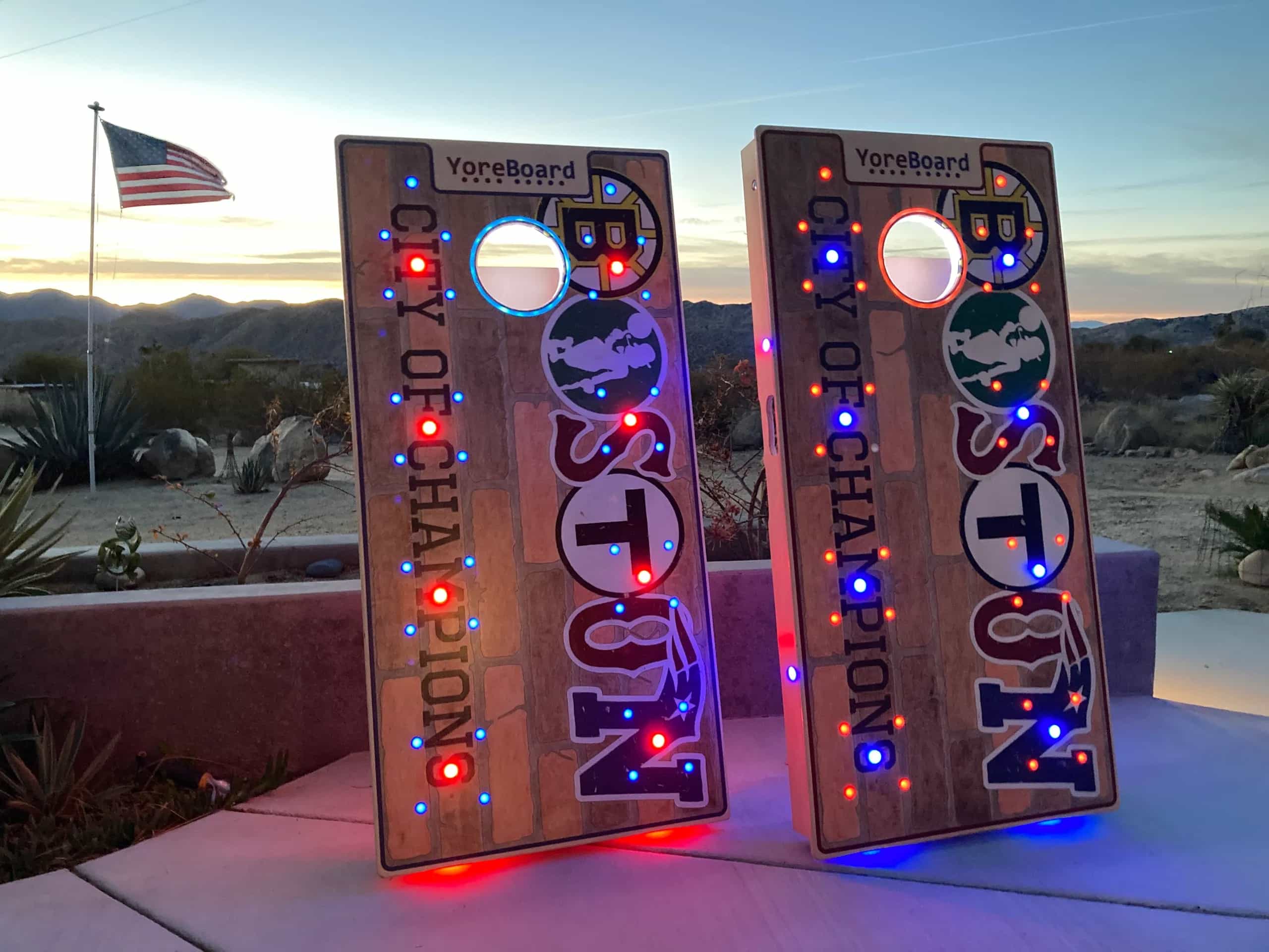 Personalized YoreBoard cornhole boards celebrating Boston's 'City of Champions' with integrated team logos and vibrant red and blue Scoring LEDs for fans.