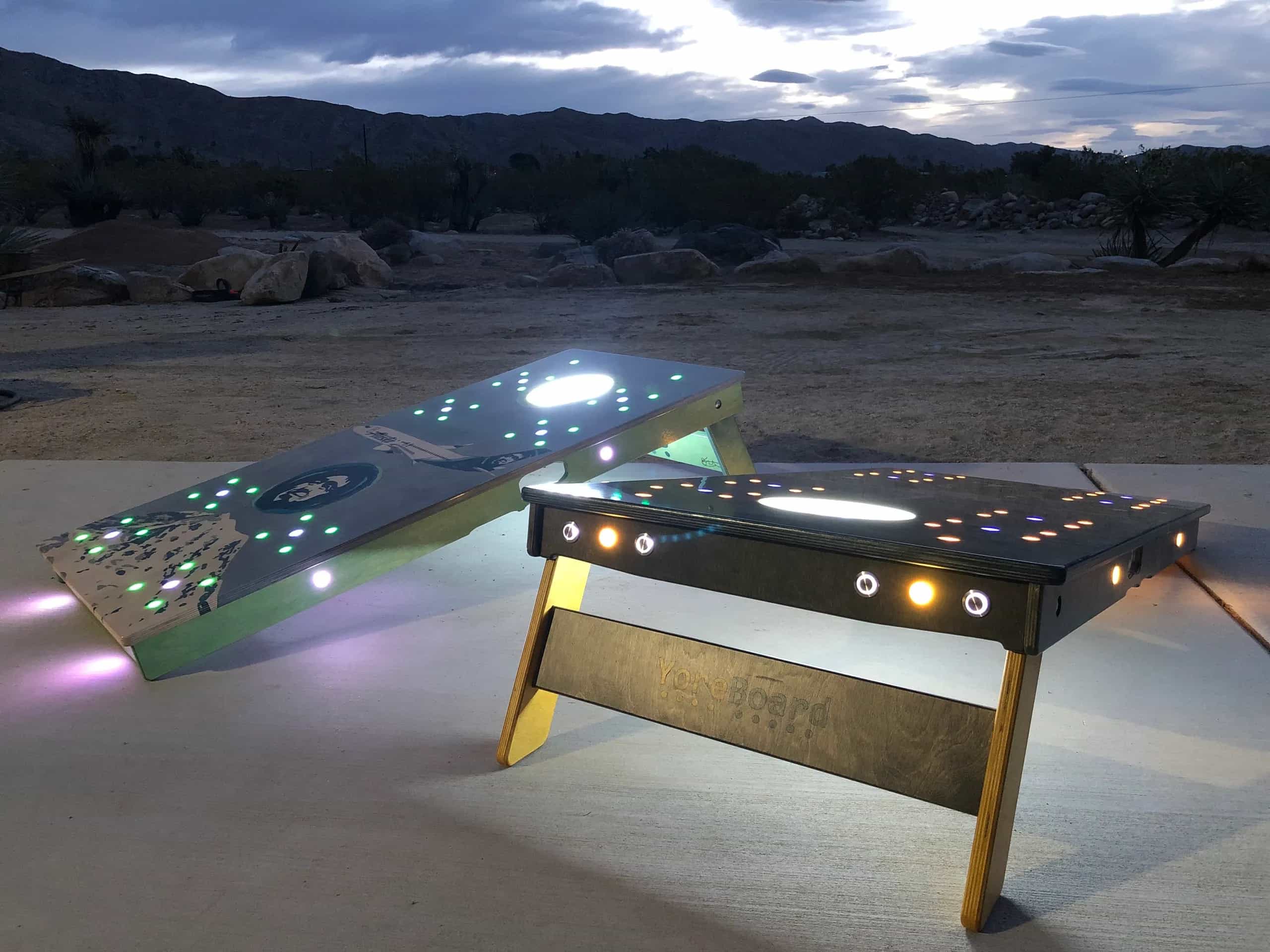 Two custom YoreBoard cornhole boards, one light-colored and one dark-colored, both featuring embedded LED lights and prominent illuminated buttons for scorekeeping, set outdoors at twilight with a desert landscape in the background.