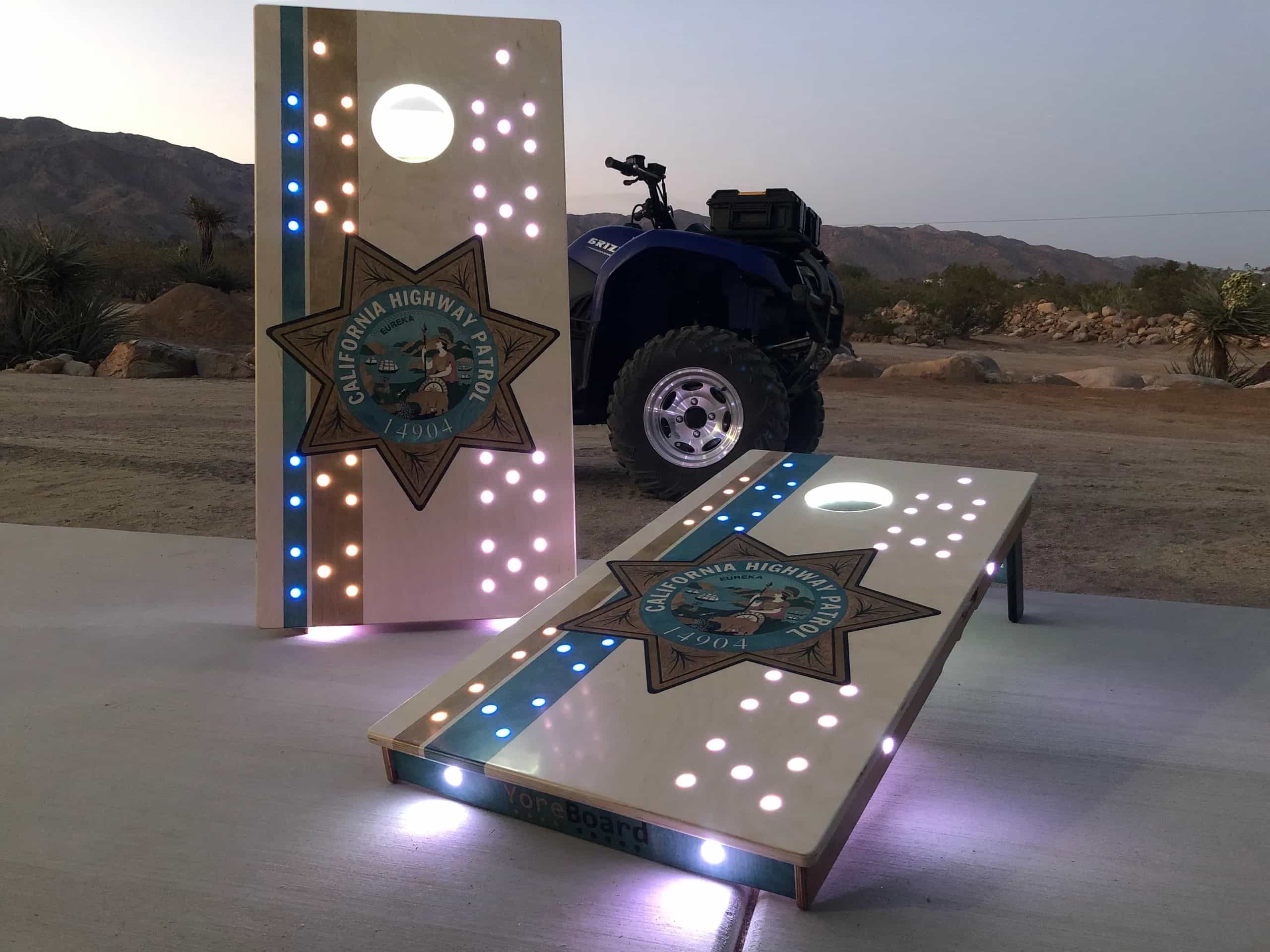 Two custom YoreBoard cornhole boards featuring the California Highway Patrol star emblem and glowing white and blue LED lights, set outdoors at twilight with an ATV.