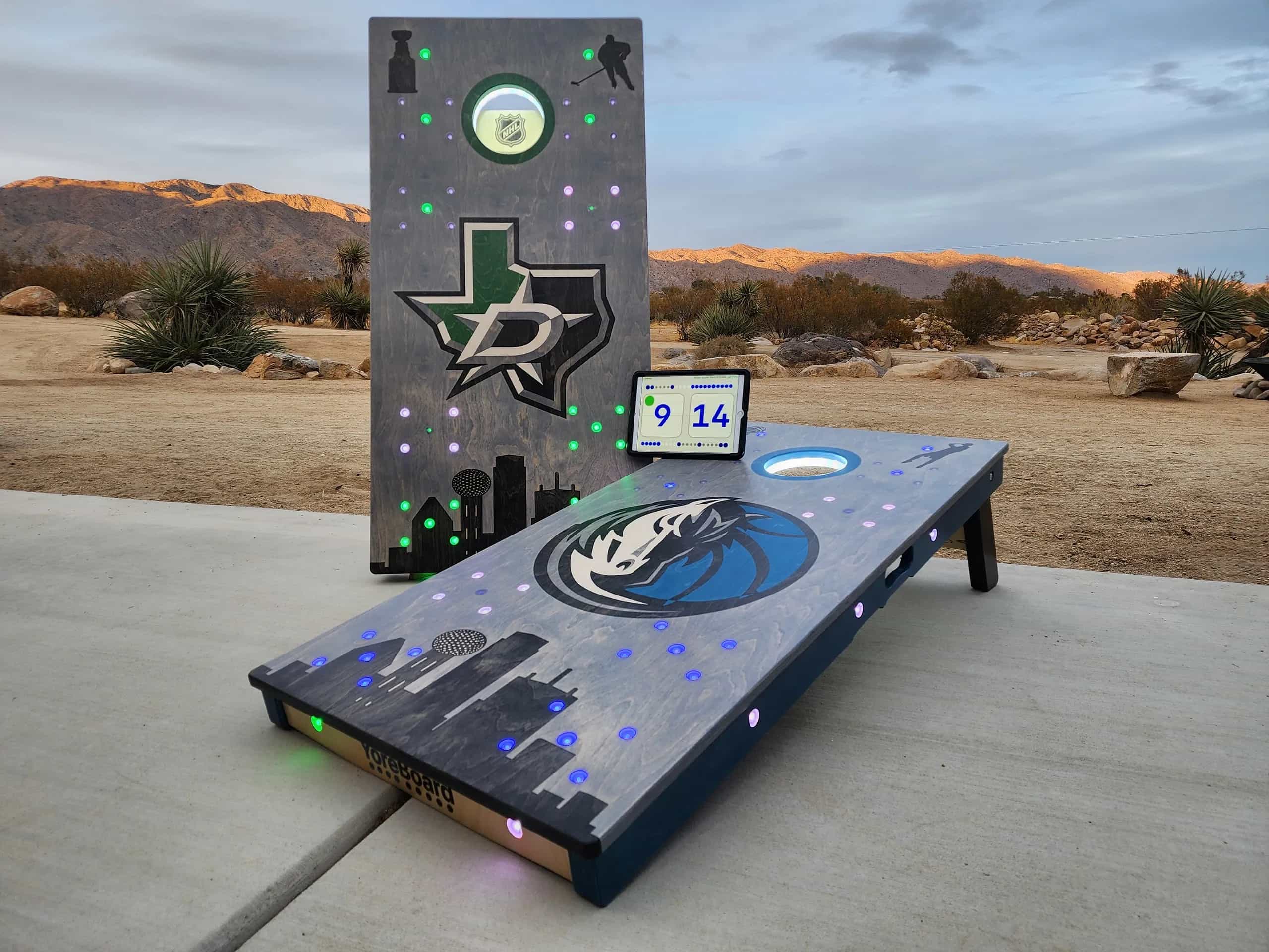 A pair of YoreBoard cornhole boards designed with Dallas sports team themes and glowing LED lights for score tracking, displayed at dusk with a portable digital scoreboard.