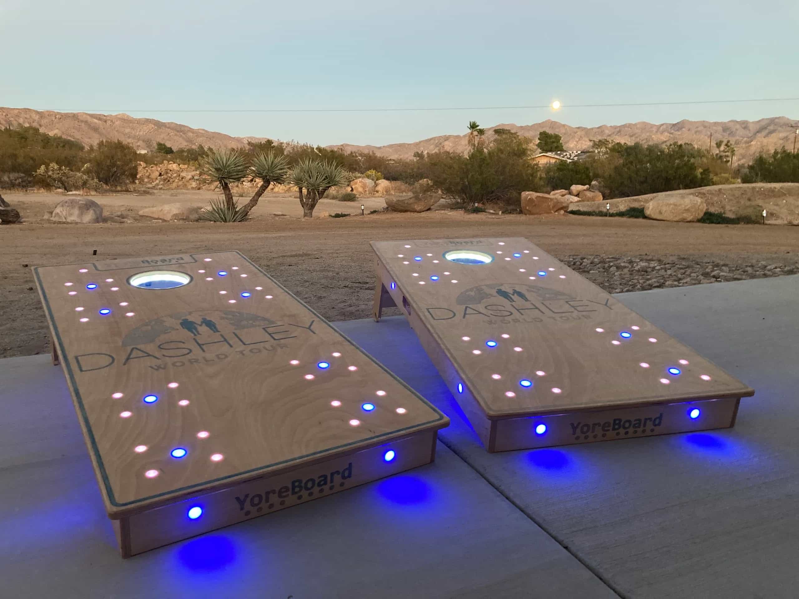 Two custom YoreBoard cornhole boards glowing with blue and pink Scoring LEDs and under-board lighting, featuring 'Dashley World Tour' art in a twilight desert setting.
