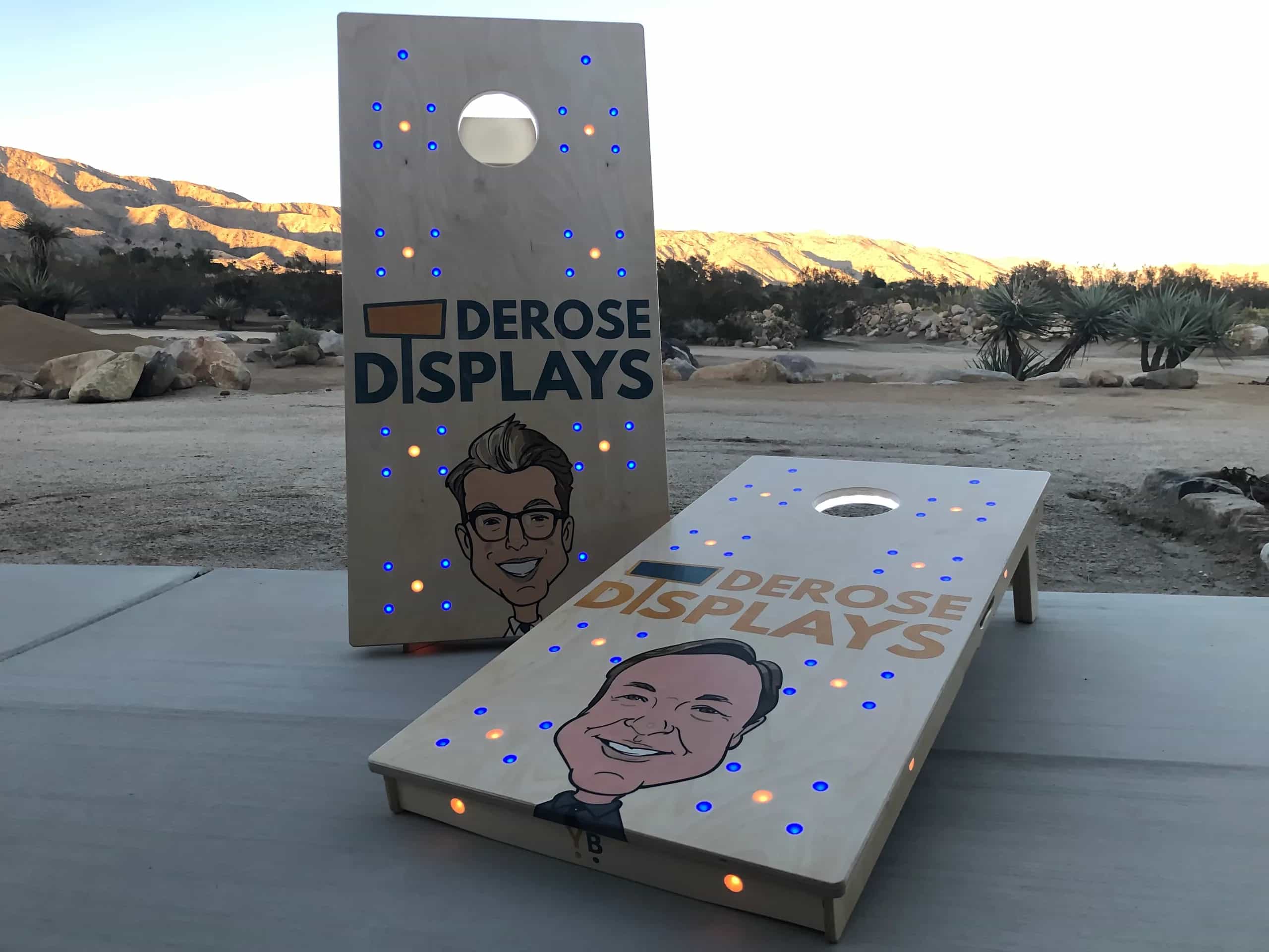 Custom YoreBoard cornhole set for Derose Displays, featuring company logo, cartoon caricatures, and blue and orange Scoring LEDs.