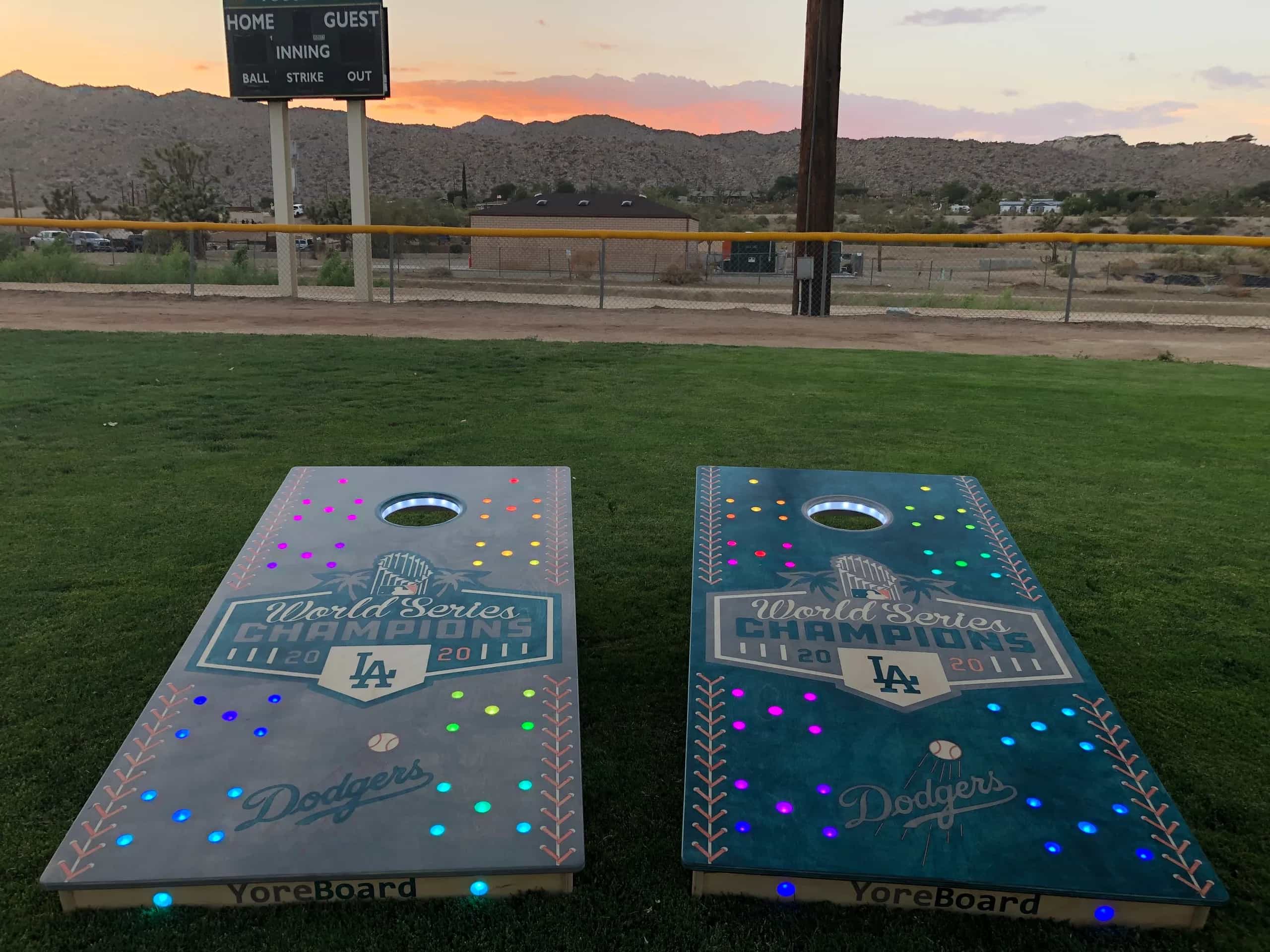 Two custom YoreBoard cornhole boards, one blue and one gray, both featuring 'World Series Champions' and Dodgers logos with embedded multi-colored LED lights, set on a grassy field in front of a traditional baseball scoreboard at sunset.