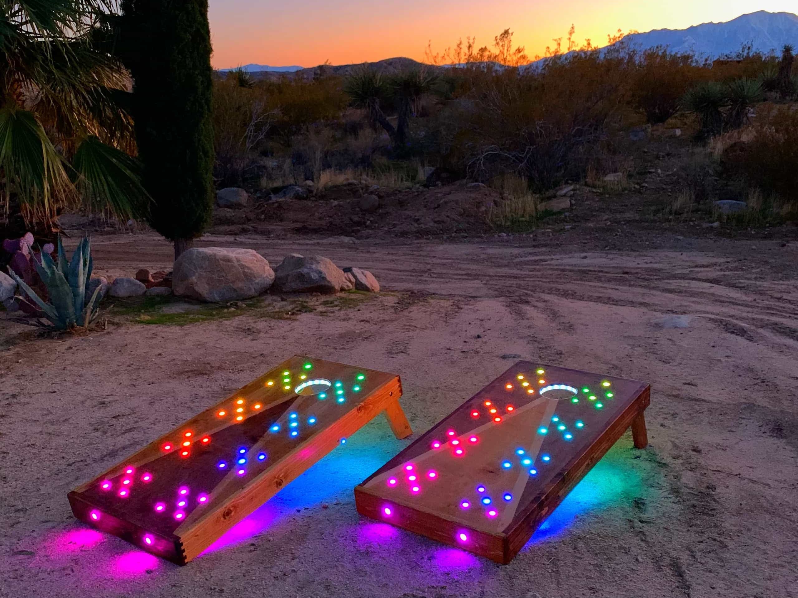 Two custom YoreBoard cornhole boards, illuminated with vibrant multi-colored LED lights, set up on a dirt path in a desert landscape with mountains and a colorful sunset in the background.