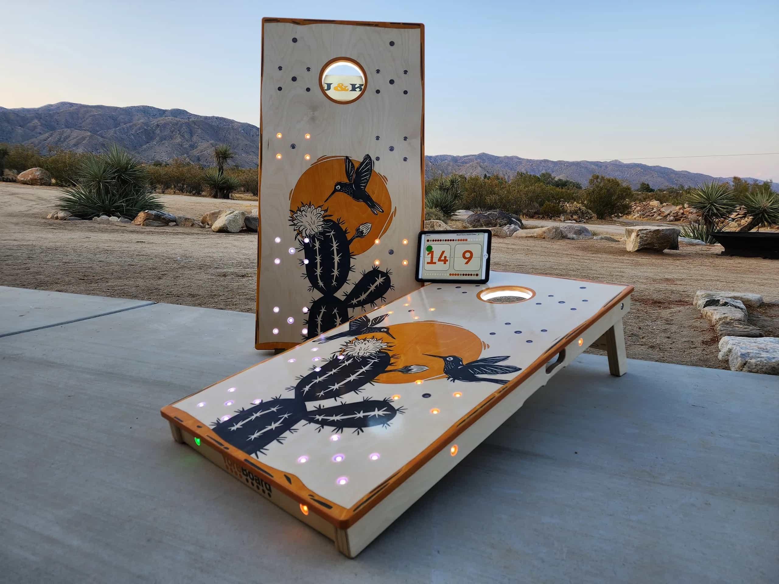 Personalized YoreBoard cornhole boards showcasing detailed hummingbird and cactus desert artwork, enhanced by glowing Scoring LEDs and an active digital scoreboard.