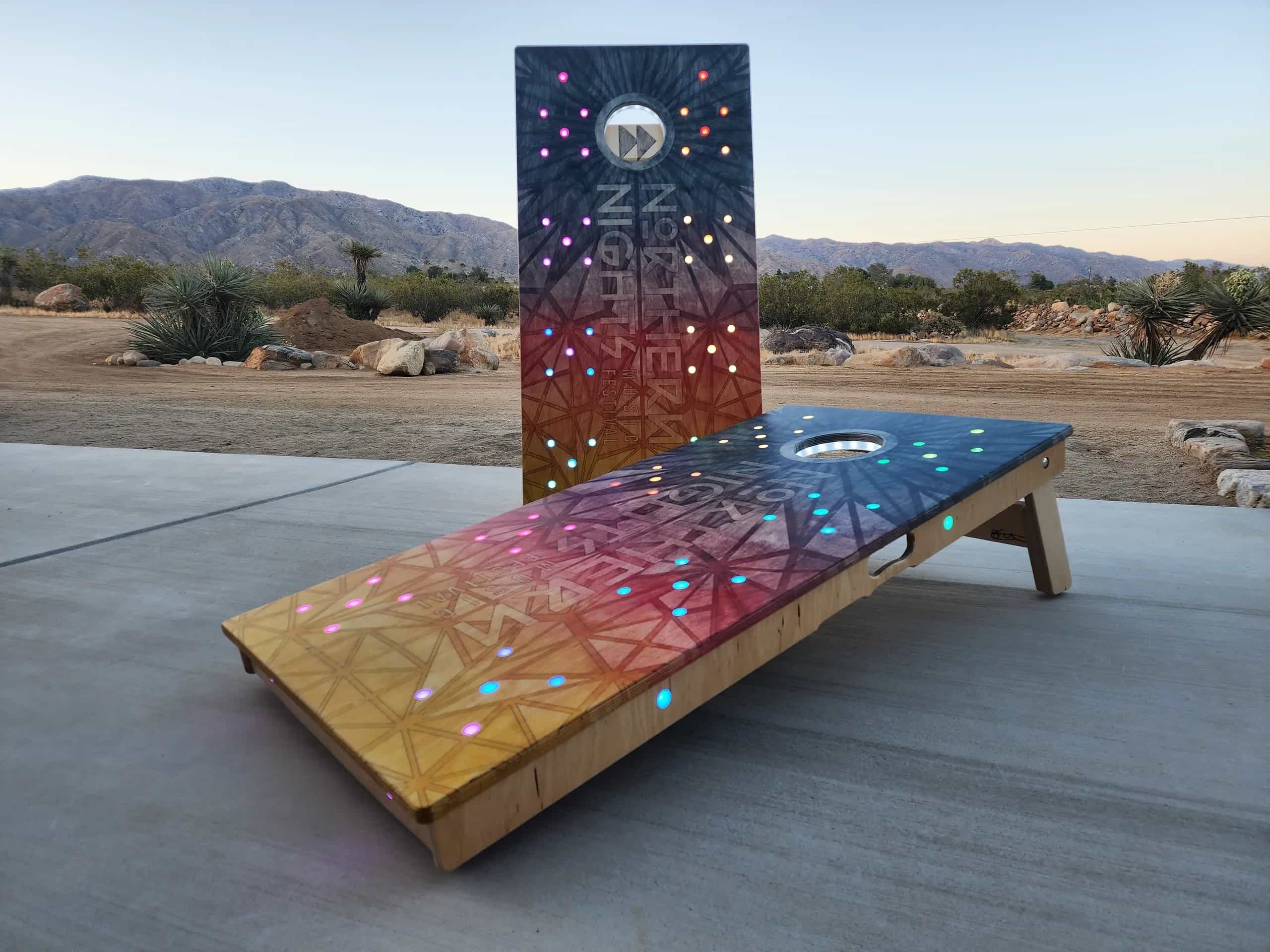 Custom YoreBoard cornhole set with a vibrant Northern Lights gradient design, geometric patterns, and multi-colored Scoring LEDs, set outdoors at twilight.