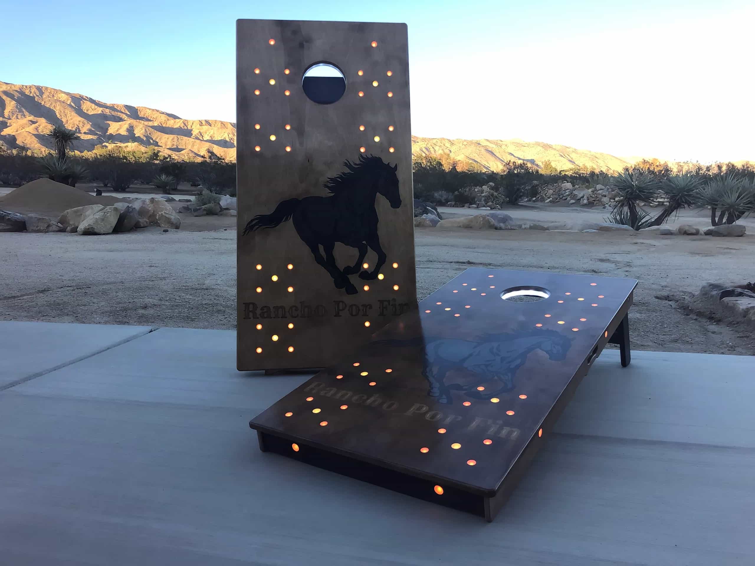 Custom YoreBoard cornhole set for 'Rancho Por Fin', featuring galloping horse silhouettes and warm orange Scoring LEDs, set outdoors at dusk.