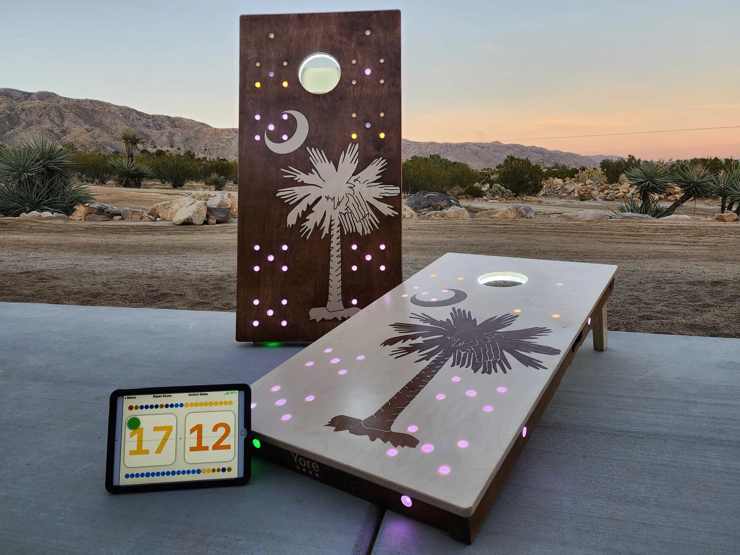 Two custom YoreBoard cornhole boards, featuring illuminated palm tree and crescent moon designs with embedded LEDs for scorekeeping, wirelessly synced via Bluetooth to the digital scoreboard displayed on a tablet.