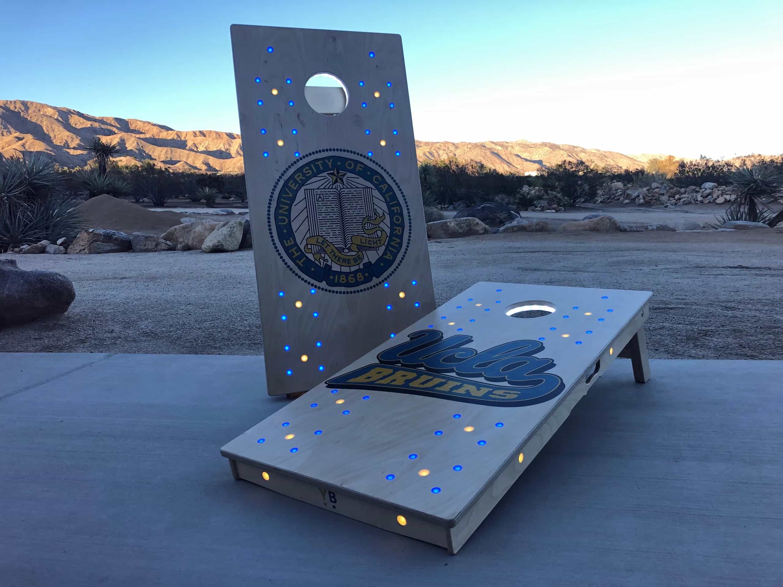 Two custom YoreBoard cornhole boards celebrating UCLA Bruins with university emblems and glowing blue and gold Scoring LEDs, perfect for tailgating.