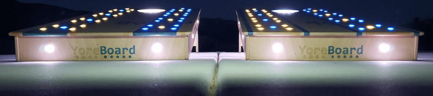 Dramatic close-up of two YoreBoard cornhole boards at night, showcasing glowing 'YoreBoard' logos, under-board lighting, and rows of blue and yellow Scoring LEDs.