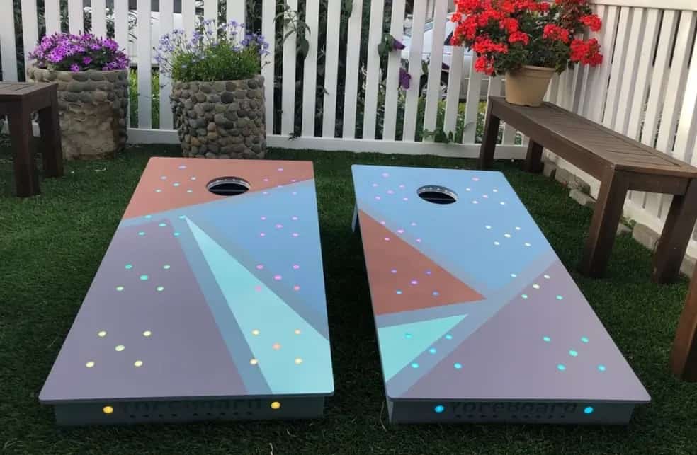Two YoreBoard cornhole boards with a modern geometric design in blue, grey, and peach tones, illuminated by multi-colored Scoring LEDs on artificial turf.