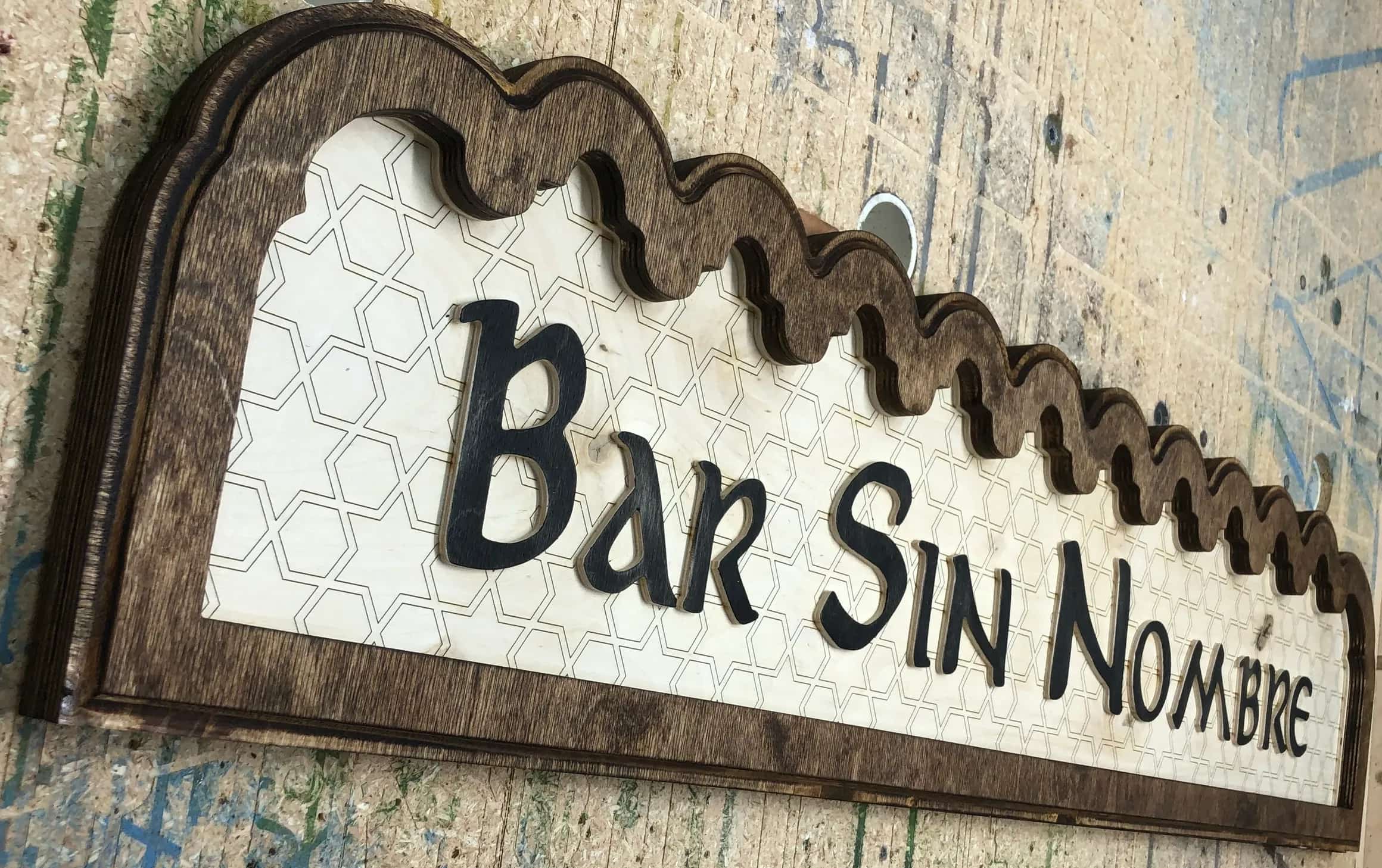 Handcrafted wooden sign for 'Bar Sin Nombre' featuring dimensional dark brown lettering. The light-colored base wood has a faint hexagonal pattern, and the sign is framed by an ornate, wavy dark wood border.