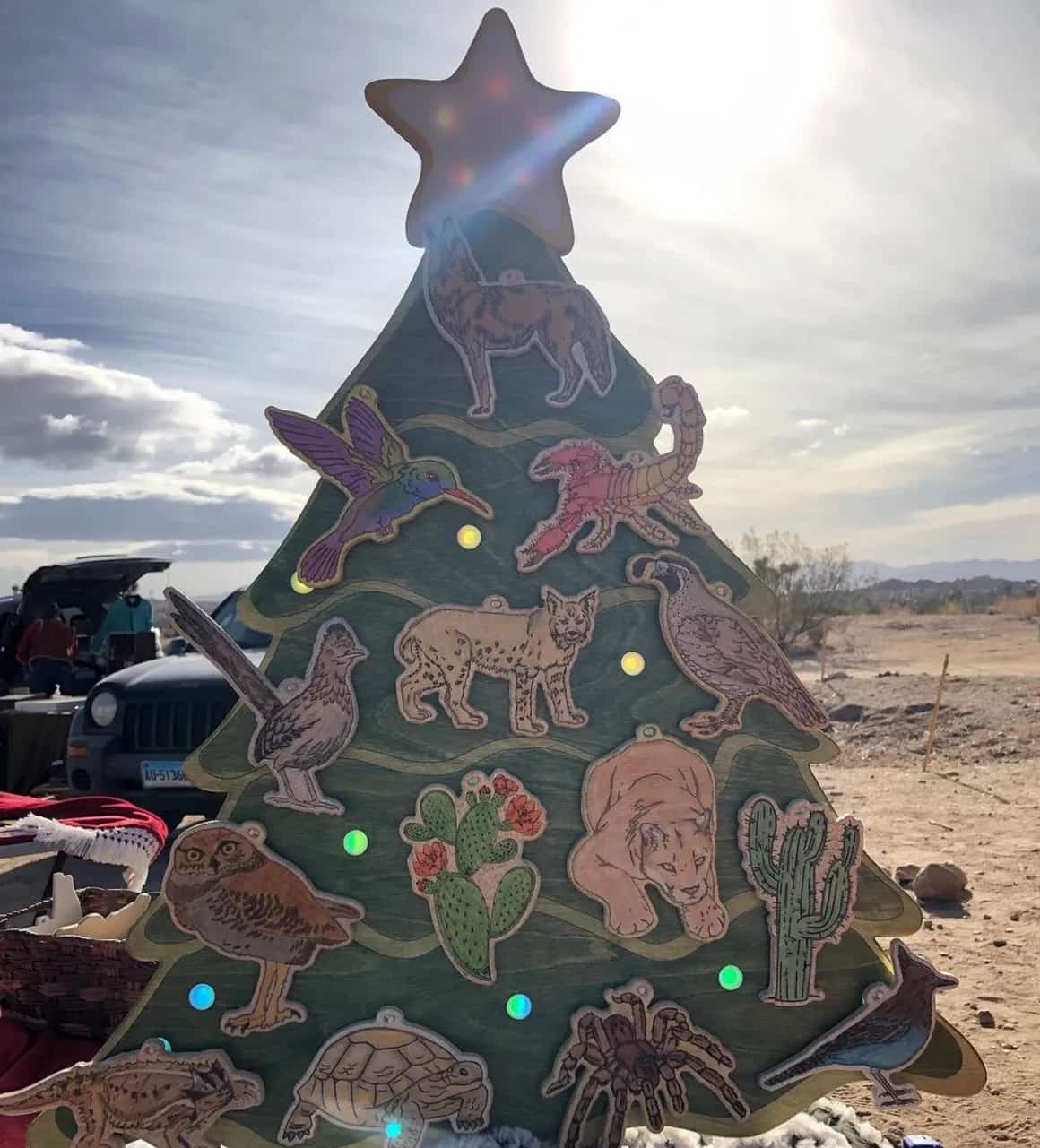 Unique holiday decoration: a green wooden Christmas tree featuring a star topper and numerous light-colored cut-out ornaments depicting desert flora and fauna such as an owl, joshua tree, and roadrunner, with sunlight in the background.