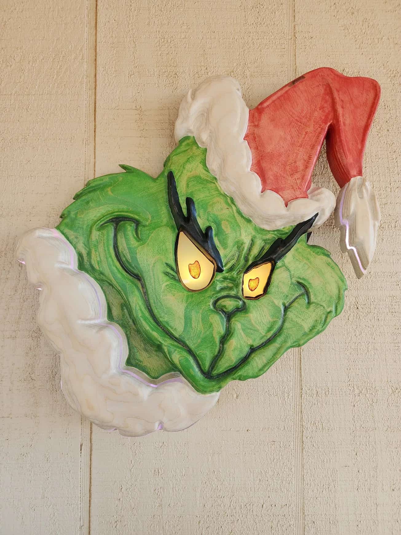 Festive holiday decoration: a handcrafted Grinch head sign with bright, illuminated eyes and a Santa hat, showcasing the character's iconic scowl and green complexion.