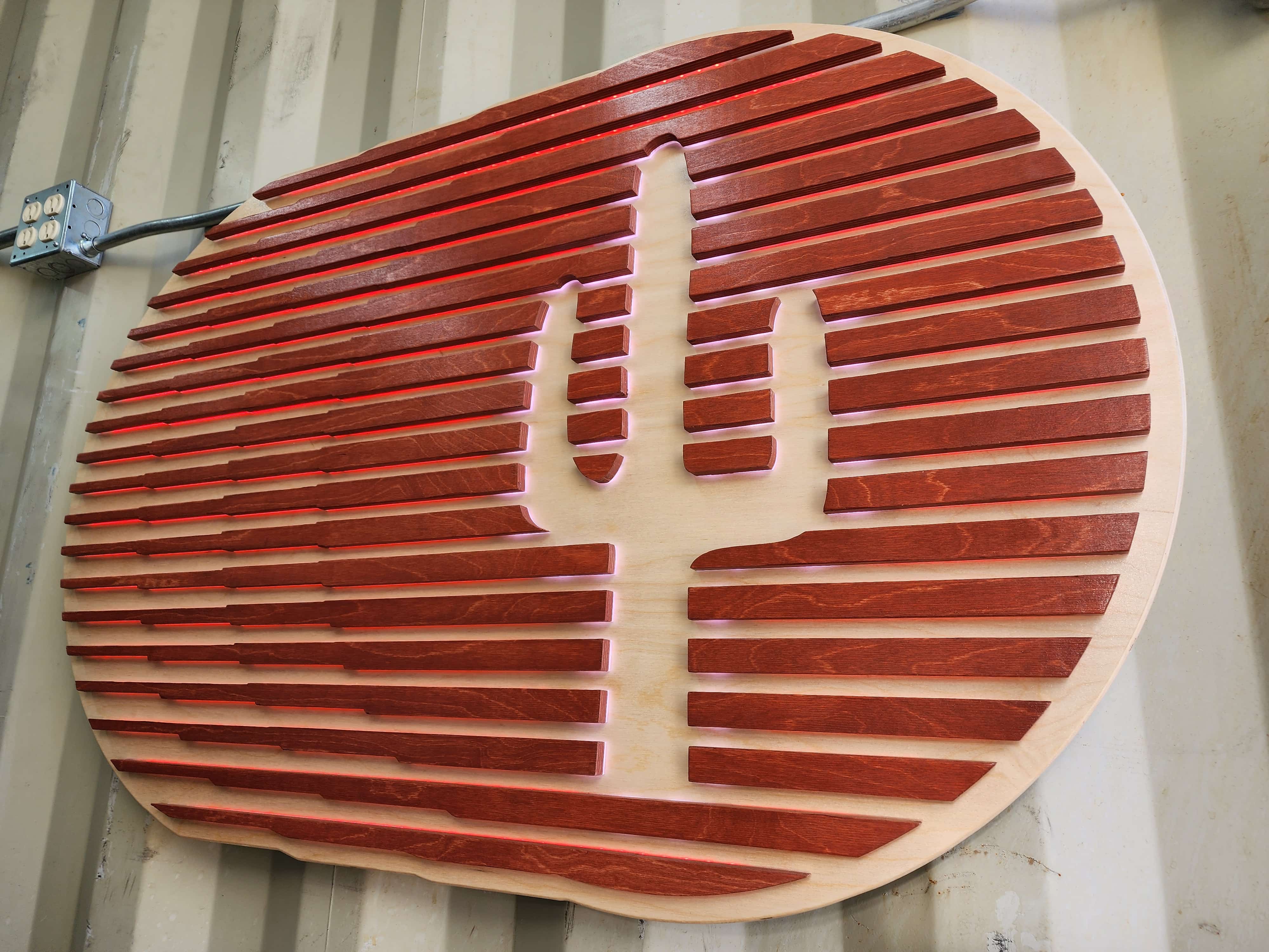 Handcrafted desert-themed sign: a saguaro cactus silhouette carved into a light wood panel, overlaid with dark wood horizontal slats. The cactus is illuminated from behind, creating a striking visual effect.
