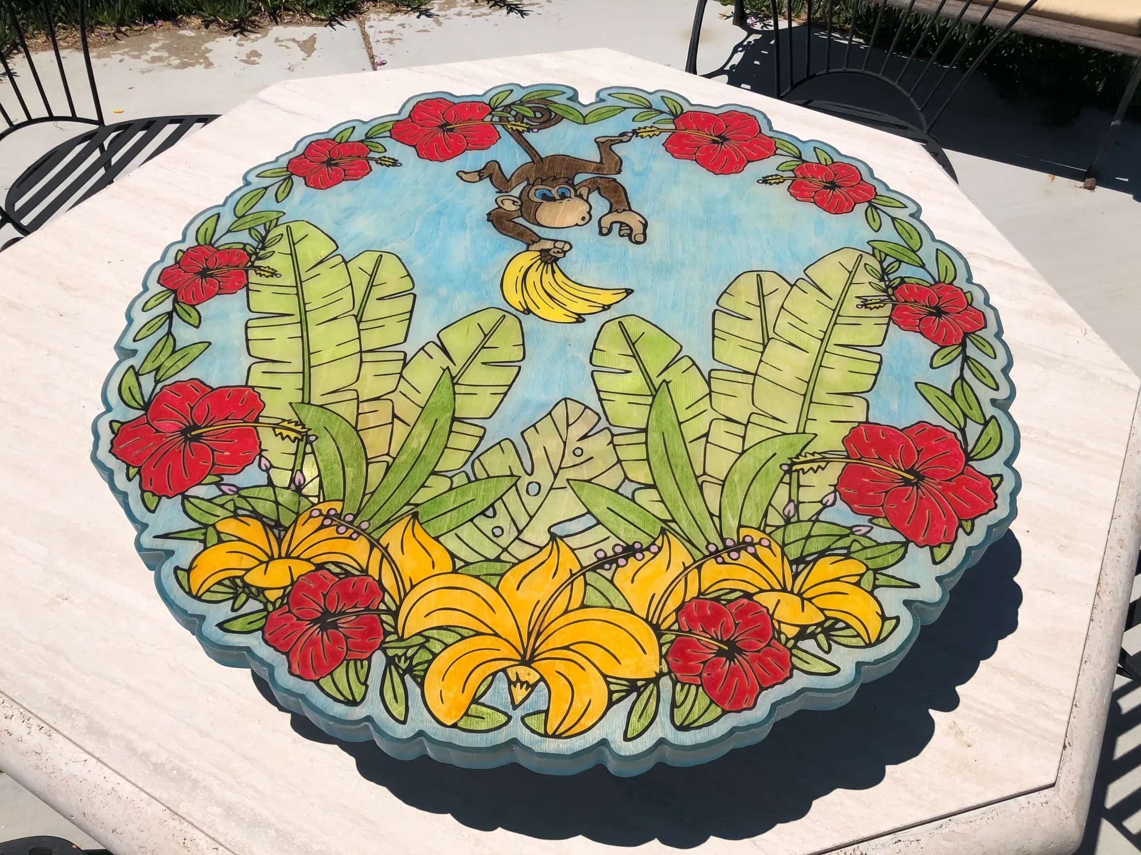 Handcrafted outdoor table surface decorated with a whimsical jungle motif. The design centers on a playful monkey, large tropical leaves, and colorful hibiscus and lily-like flowers, all painted in vivid hues.