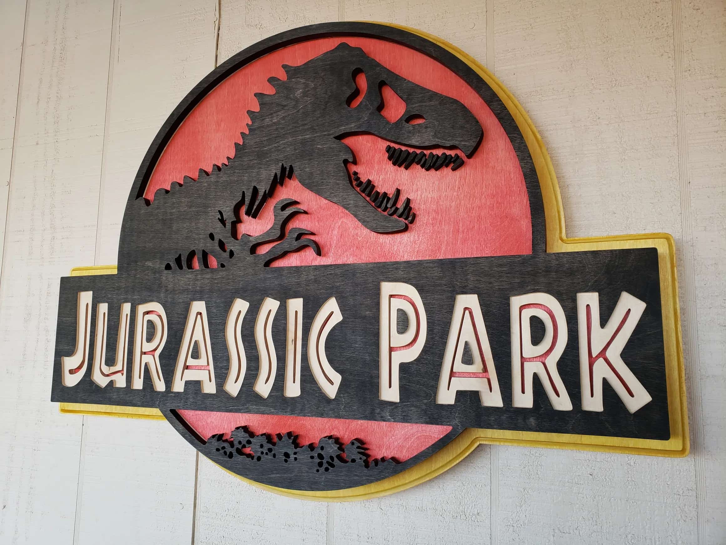 Handcrafted Jurassic Park emblem: a dimensional wooden sign featuring the classic T-Rex and title, with contrasting wood layers in black, red, and natural tones, creating a striking visual effect.