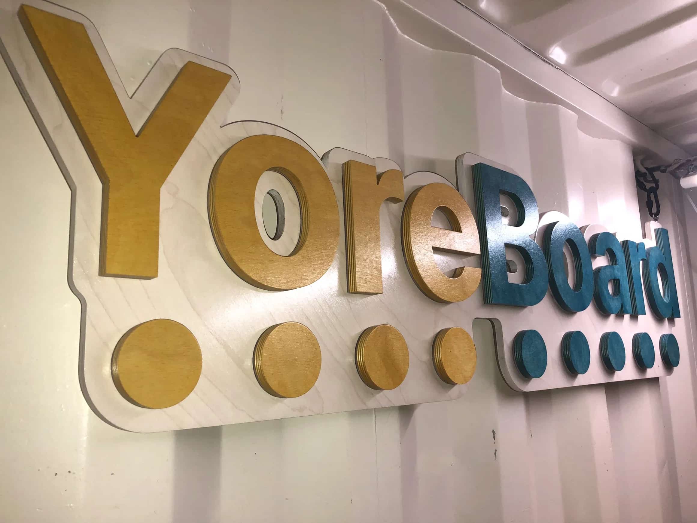 Handcrafted YoreBoard brand sign: a multi-layered wooden display with the company name in two distinct wood finishes and a row of circular accents, mounted on a textured industrial wall.