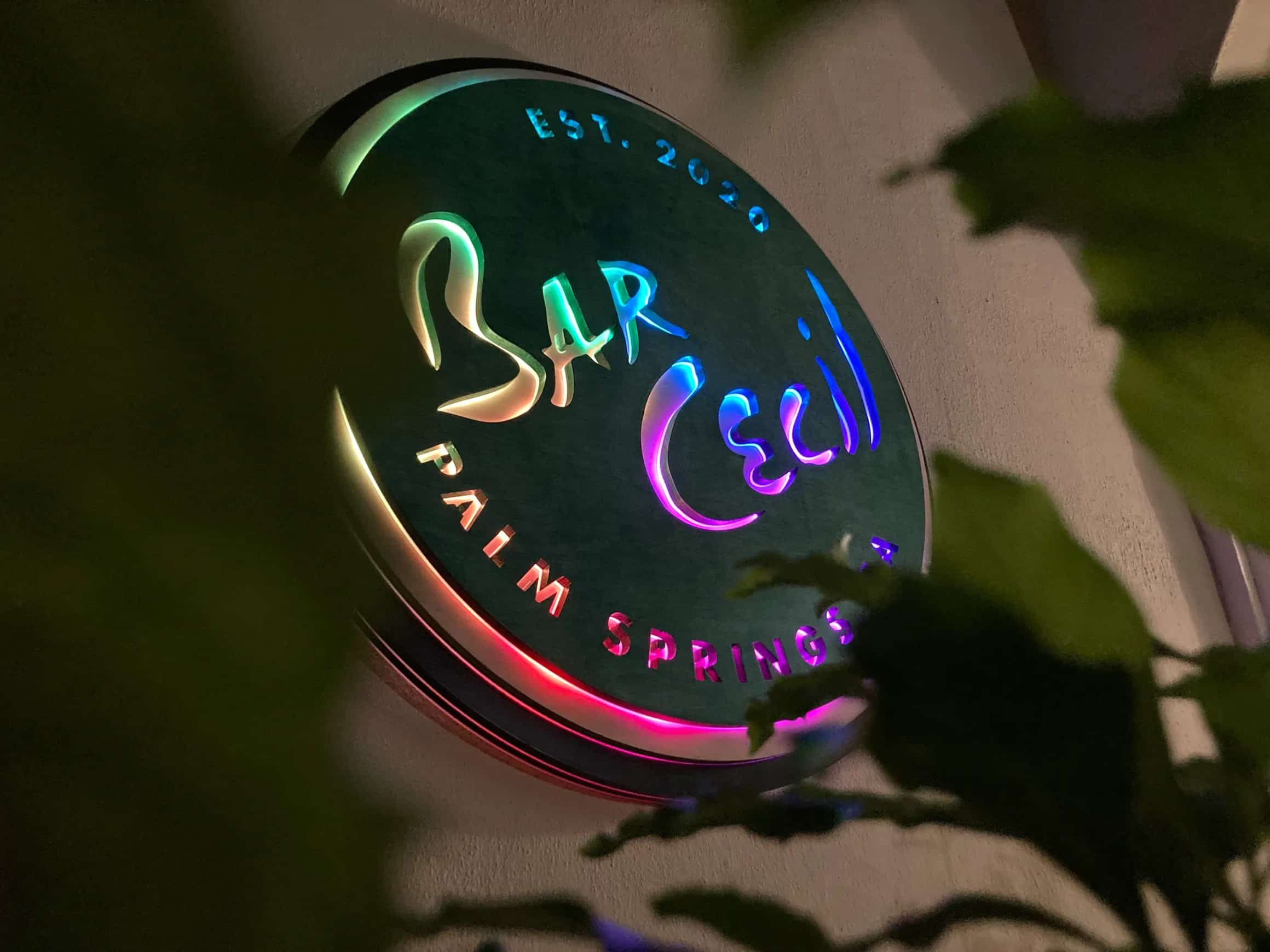 Nighttime view of the Bar Cecil restaurant sign, featuring its distinctive circular logo with 'Bar Cecil' and 'Palm Springs, CA Est. 2020' text. The sign is brightly lit with a changing spectrum of colors, partially visible behind dark leaves.