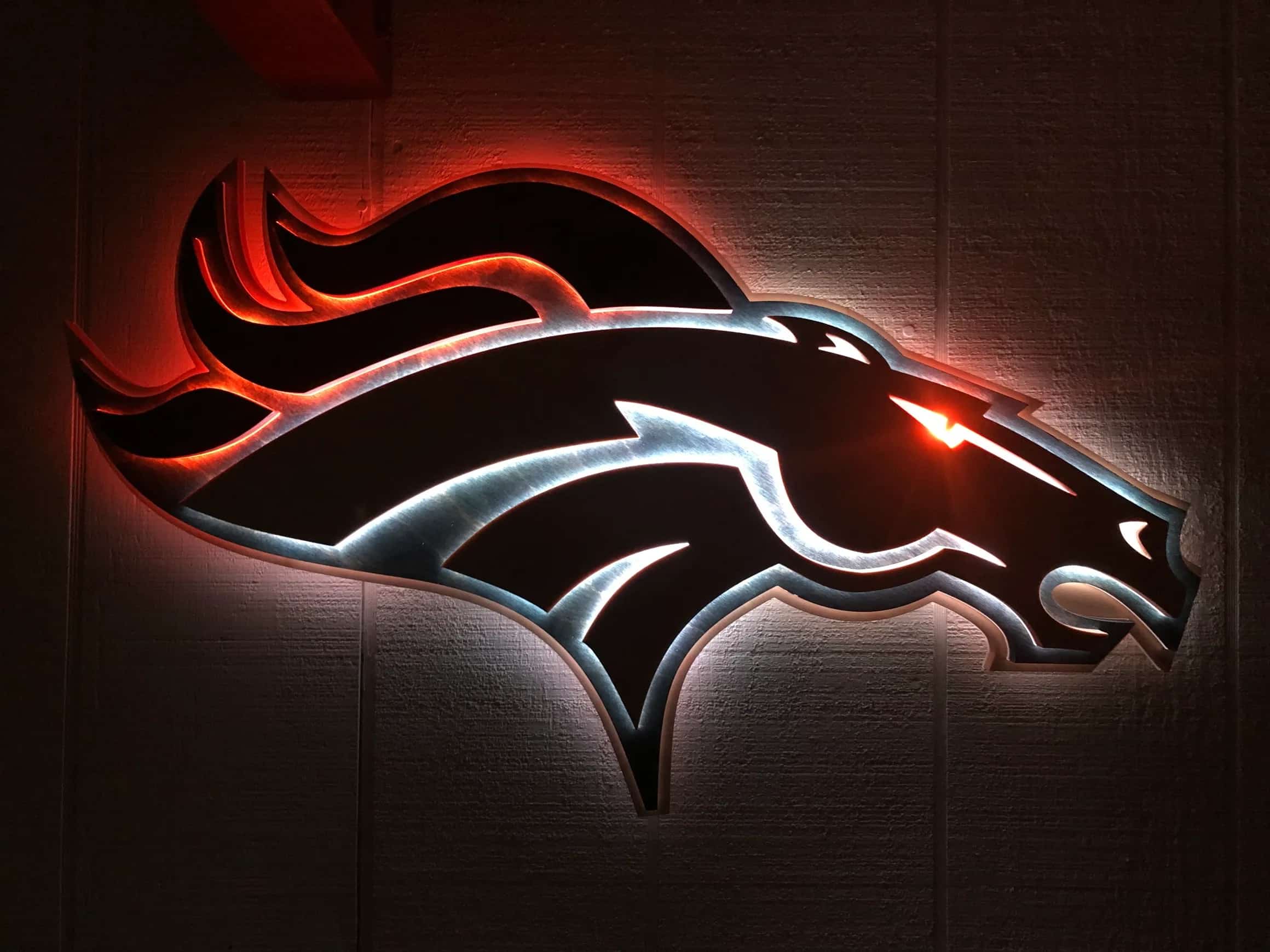 Custom-made, multi-layered Denver Broncos logo sign, showcasing the horse head design with vibrant LED backlighting. The mane glows orange, while the lower outline emits a cool white or light blue, creating a dramatic effect against a dark background.