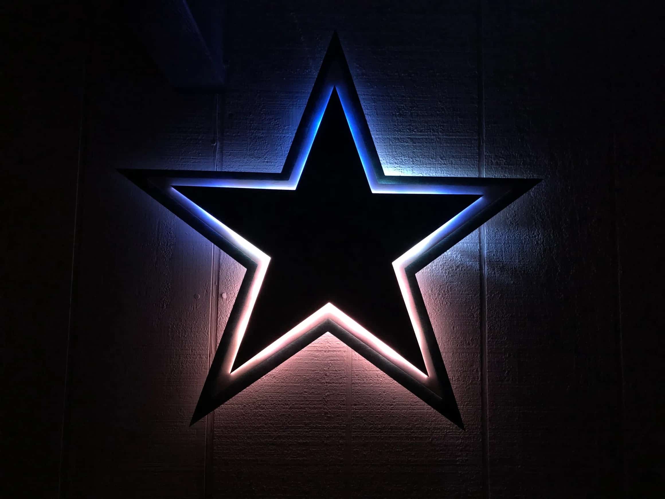 Illuminated Dallas Cowboys star logo, with the top half glowing blue and the bottom half glowing white, creating a gradient effect. The star has a dark center and is mounted on a textured wall in a dark setting.
