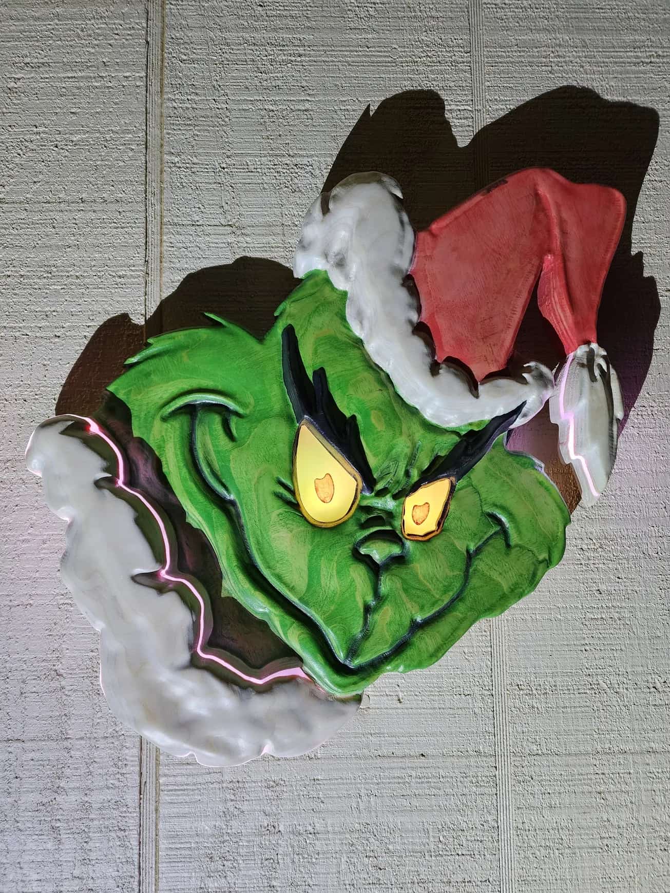 Custom-made, dimensional sign of the Grinch, featuring his signature green face, mischievous yellow glowing eyes, and a classic red Santa hat. The outline of his fur is subtly lit with white LEDs, adding depth to the holiday decoration.