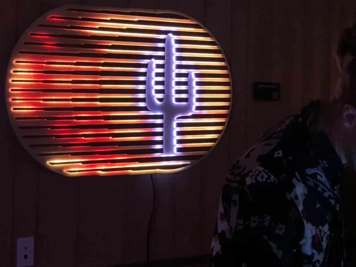 Handcrafted desert-themed sign: a saguaro cactus silhouette illuminated with cool tones, set against a background of horizontal slats glowing with warm red and orange light. The sign creates a vibrant, contrasting glow in a dimly lit dance floor in front of the DJ.