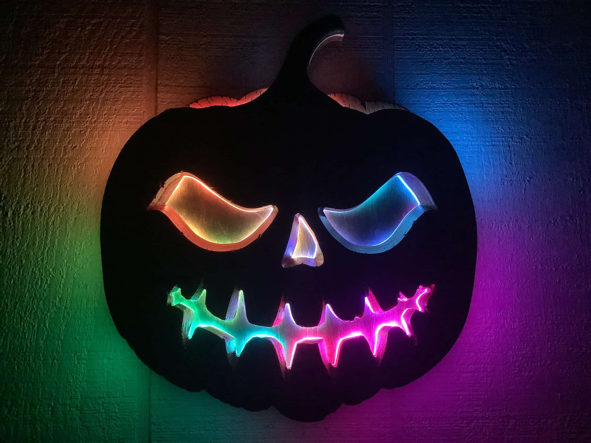 Custom-made, multi-layered jack-o'-lantern sign with a carved face that glows with a vibrant spectrum of LED colors. Each eye and section of the jagged mouth displays a different hue, creating a dynamic and spooky Halloween decoration.